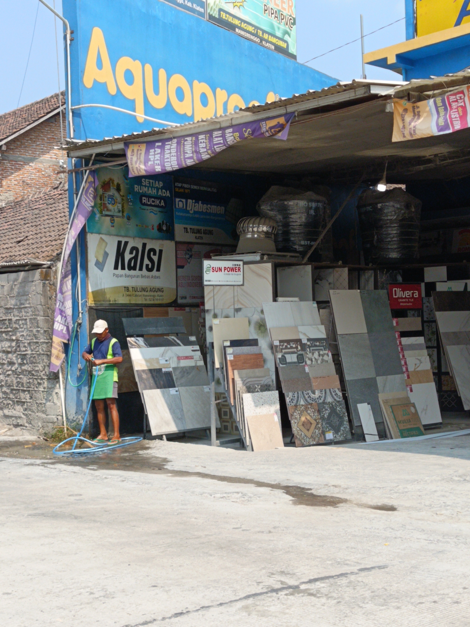 A medium shot captures a man hosing down the concrete ground in front of a building that appears to be a store or warehouse. The man, wearing a blue shirt, a green apron, and a white cap, holds a blue hose that sprays water onto the ground. He is standing near a display of various tiles. Behind him, a blue building with yellow lettering that reads "Aquapro" is visible, along with other signage and banners, including one for "Kalsi" and another with "Djabesmen". The scene is outdoors with a bright sky and shadows cast by the structures. The location is identified as Manisrenggo, Indonesia.