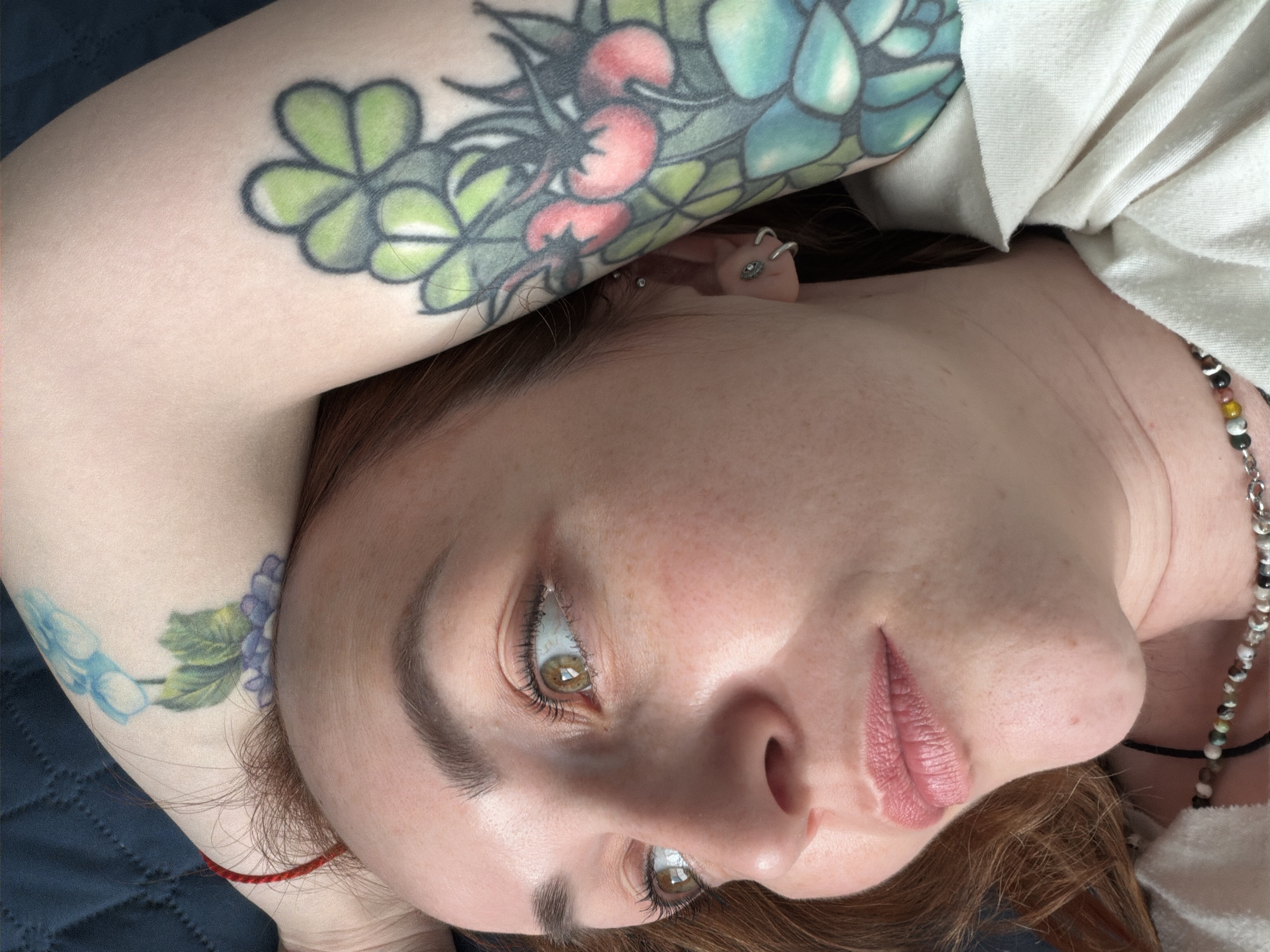 A woman with freckled skin and brown eyes is lying on a blue quilted surface. Her right arm is bent and raised, with a tattoo of clovers, red berries, and teal succulents visible on her upper arm. Her left arm is also bent and raised, showing a tattoo of blue flowers on her inner arm. She has multiple piercings on her left ear. She is wearing a light-colored shirt and a beaded necklace. A red string bracelet is visible on her left wrist. The location cannot be determined from the image.