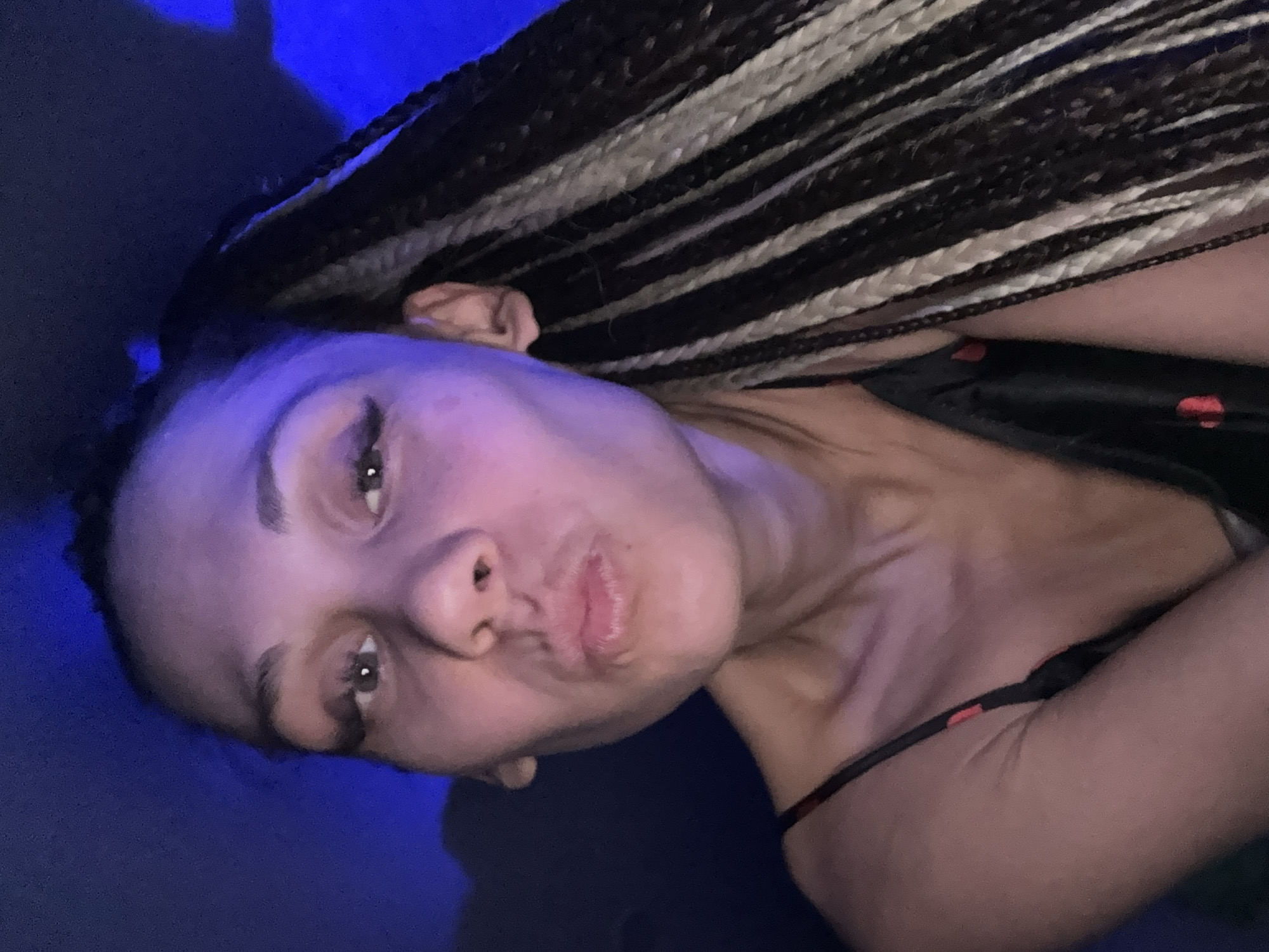 A woman is posing for a selfie. The background is a blue hue. The woman is wearing a black and red top and has black and white braids. She is pouting her lips. Location unknown.