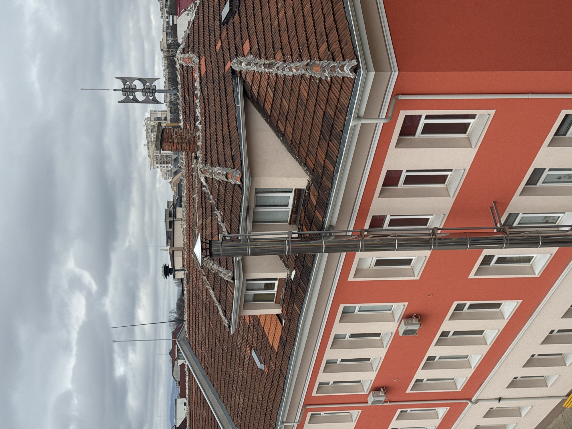 The image depicts a building in Sofia, Bulgaria, characterized by its red and white facade and brown tiled roof. The building has multiple rectangular windows with light-colored frames. Air conditioning units are attached to the exterior wall. A prominent feature is the large siren mounted on a rooftop in the background, with a cloudy sky overhead. Chimneys are also visible on the roofs. Metallic pipes run alongside the building's exterior.