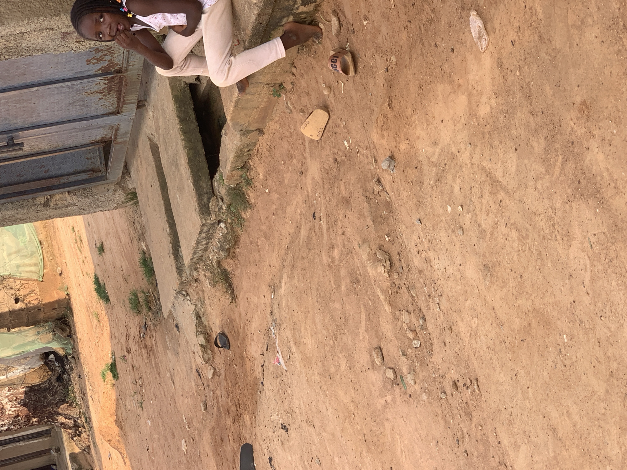 In Sabon Tasha, Nigeria, a young girl with braided hair and wearing a white tank top with pink trim and light-colored pants sits on a concrete ledge next to a rusty metal gate. She is biting her fingernails. Scattered on the dusty, dirt ground are several pairs of sandals, including a black flip-flop, two tan slides, and a brown slide with "GG" logo. A discarded clear plastic bottle is also visible on the ground. The environment appears to be an outdoor urban setting with a large tree and some overgrowth in the background.
