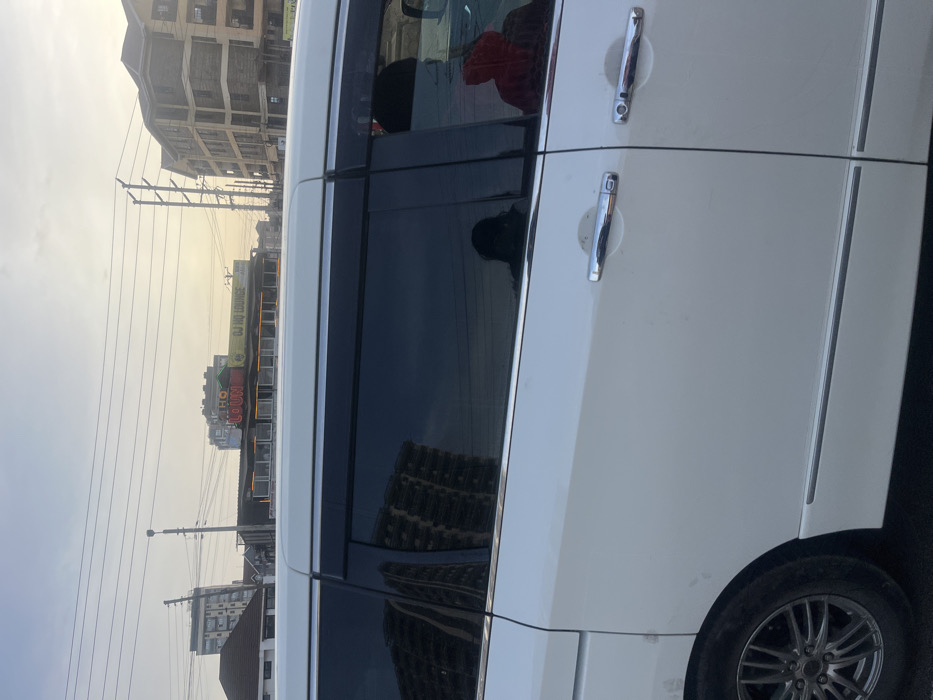 The image captures a street scene in Gitothua ward, Kenya, featuring a white vehicle on the right and an urban landscape in the background. The vehicle, likely a minivan, shows part of its side body, chrome door handles, dark tinted windows, and a multi-spoke wheel. Reflections are noticeable in the vehicle's windows; a person wearing a red top is faintly visible inside, reflected in the rear window, while a more indistinct figure is seen in the side window. A building is also visibly reflected in the tinted glass.
The background reveals a street lined with buildings. Overhead, numerous power lines crisscross the sky, supported by utility poles. The sky appears hazy with a yellowish hue, suggesting either early morning or late afternoon. On a building in the mid-ground, bright signs are visible, displaying the text "DJ HO LOUNGE" and "HOURI," indicating a commercial establishment, possibly a bar or entertainment venue. The overall setting is an urban or semi-urban environment.