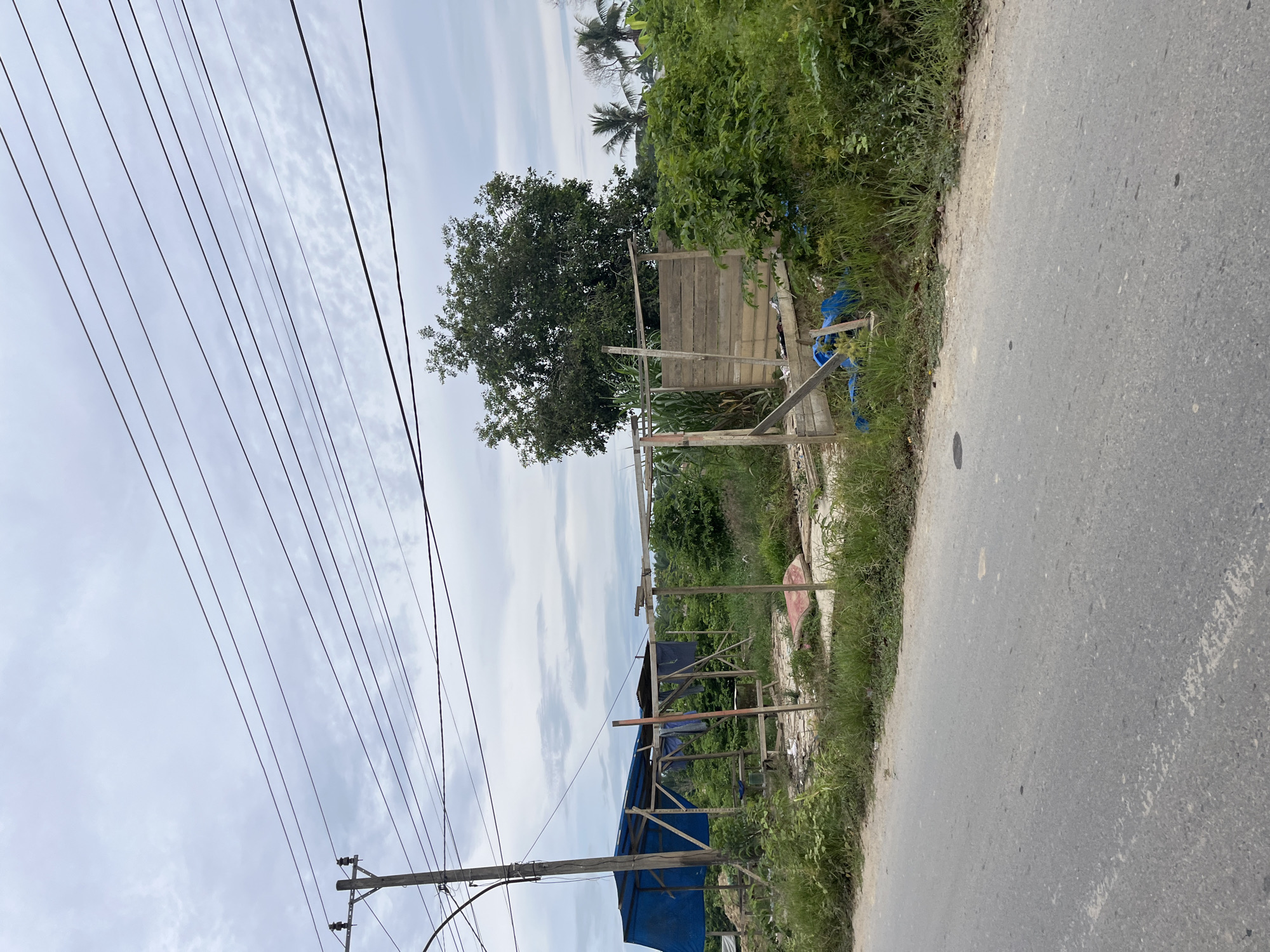 The image captures a roadside scene in Bundar, Indonesia. The foreground features an asphalt road with a white painted line. Beyond the road, a patch of overgrown grass and vegetation borders an area with remnants of a makeshift structure. This structure appears to be a partially dismantled stall or shelter, with wooden posts and a blue tarp visible. Scattered debris, including what looks like a mattress or large cushion, blue plastic sheeting, and other refuse, lies on the ground within the vicinity of the structure. In the background, a large tree stands prominently, with more greenery and palm trees visible further in the distance. Utility poles and overhead power lines traverse the sky, which is overcast with gray clouds. The overall impression is one of a rural or semi-urban environment, possibly undergoing development or in a state of disrepair.