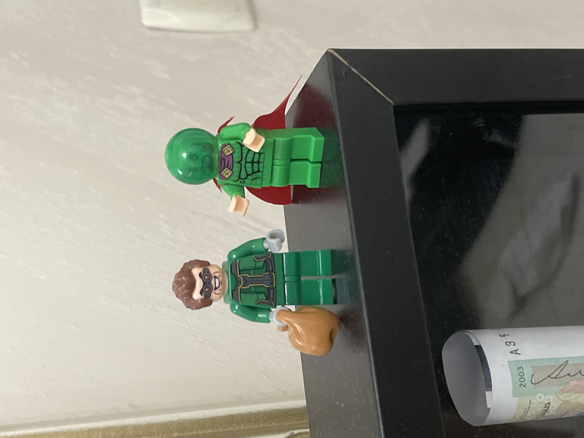 The image, rotated 90 degrees clockwise, displays two Lego minifigures positioned on a dark, possibly black, shelf or picture frame. A plain, light-colored wall with subtle texture forms the background.
The upper minifigure features a translucent green domed head and a green body, reminiscent of Mysterio. It wears a red cape, and its torso is printed with muscle detailing and a dark purple chest piece accented with gold.
The lower minifigure is also dressed in green, with a brown hairpiece and a smiling face printed with goggles or glasses. Its torso shows detailed printing in dark grey and black. This minifigure holds a light brown, irregular object, which appears to be a cooked turkey leg or similar food item.
On the right side of the dark surface, a rolled-up piece of paper, likely a banknote, is partially visible. The year "2003" and the characters "A35" are clearly discernible on the paper, alongside a signature or text in a non-Latin script, likely Cyrillic. Considering the location context of Ihtiman, Bulgaria, this is consistent with a Bulgarian lev banknote from that year. The dark display surface shows minor scuffs and dust. The scene depicts a static display, with no apparent activity or discernible emotions.