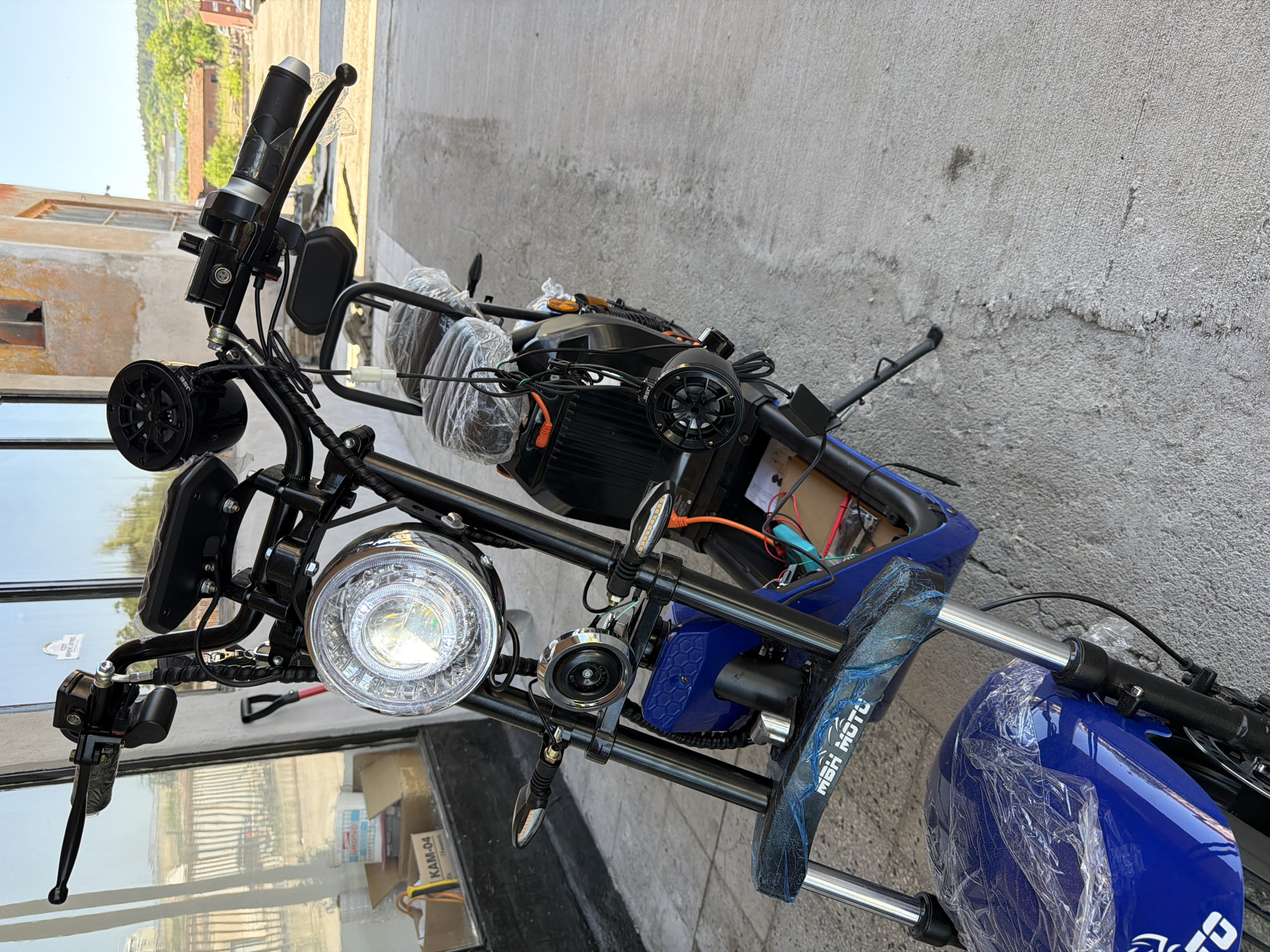 An electric scooter is parked on a concrete surface in front of a building with large windows. The scooter is blue and black with a chrome headlight and turn signals. It has two seats, both covered in plastic wrap. There are speakers mounted on the handlebars and on the back of the scooter. The building appears to be under construction or renovation, with exposed brick and peeling paint. The background shows a sunny sky and some trees.