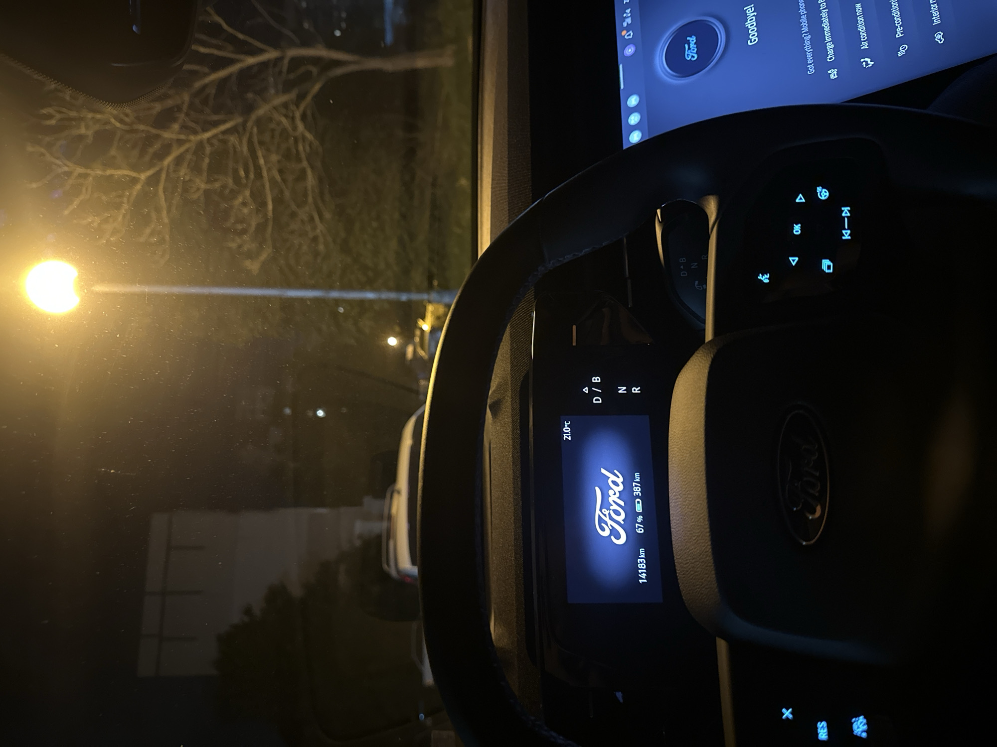 The image captures a nighttime scene from the driver's perspective inside the cabin of a Ford Mustang Mach-E, in an urban or suburban area within Sofia, Bulgaria.
In the foreground, the black leather steering wheel is prominent, featuring a subtle "Ford" logo in its center. The left spoke contains three illuminated blue buttons, including an 'X' symbol and 'RES' (likely for cruise control resume). The right spoke features illuminated blue directional arrows, an 'OK' button, a steering wheel icon, and media control buttons (skip backward, play/pause, skip forward).
Behind the steering wheel, a digital instrument cluster displays the white "Ford" logo against a dark blue background. The ambient temperature is indicated as "21.0°C". Below the logo, "14183 km" is displayed, alongside a battery icon showing "67%" charge and an estimated range of "387 km". To the right of this display, the gear selector indicator shows "D / B", "N", and "R", with "D / B" appearing active.
To the right of the instrument cluster, a large, vertical infotainment touchscreen shows a blue interface. A circular "Ford" logo is at the top, followed by the greeting "Goodbye!". Below this, a list of prompts includes "Got everything? Mobile phone...", "Charge immediately to...", "Air condition now", "Pre-condition now", and "Interior r..." (partially visible), each with an associated icon. The top right of this screen shows icons for signal strength, Wi-Fi, battery level, and a clock (specifics unreadable).
Through the windshield, the exterior scene is illuminated by a bright, warm yellow street light positioned on a pole to the upper right. A bare-branched tree stands directly behind this streetlight. On the distant left, a large, light-colored building with dark horizontal architectural features is visible. Faint reflections of the car's interior lights and minor smudges are present on the windshield. Several other vehicles are discernible in the distance, including a white SUV-like car ahead and a yellow vehicle further down the street to the right. The overall environment suggests a typical urban or suburban street or parking area. No people are visible.