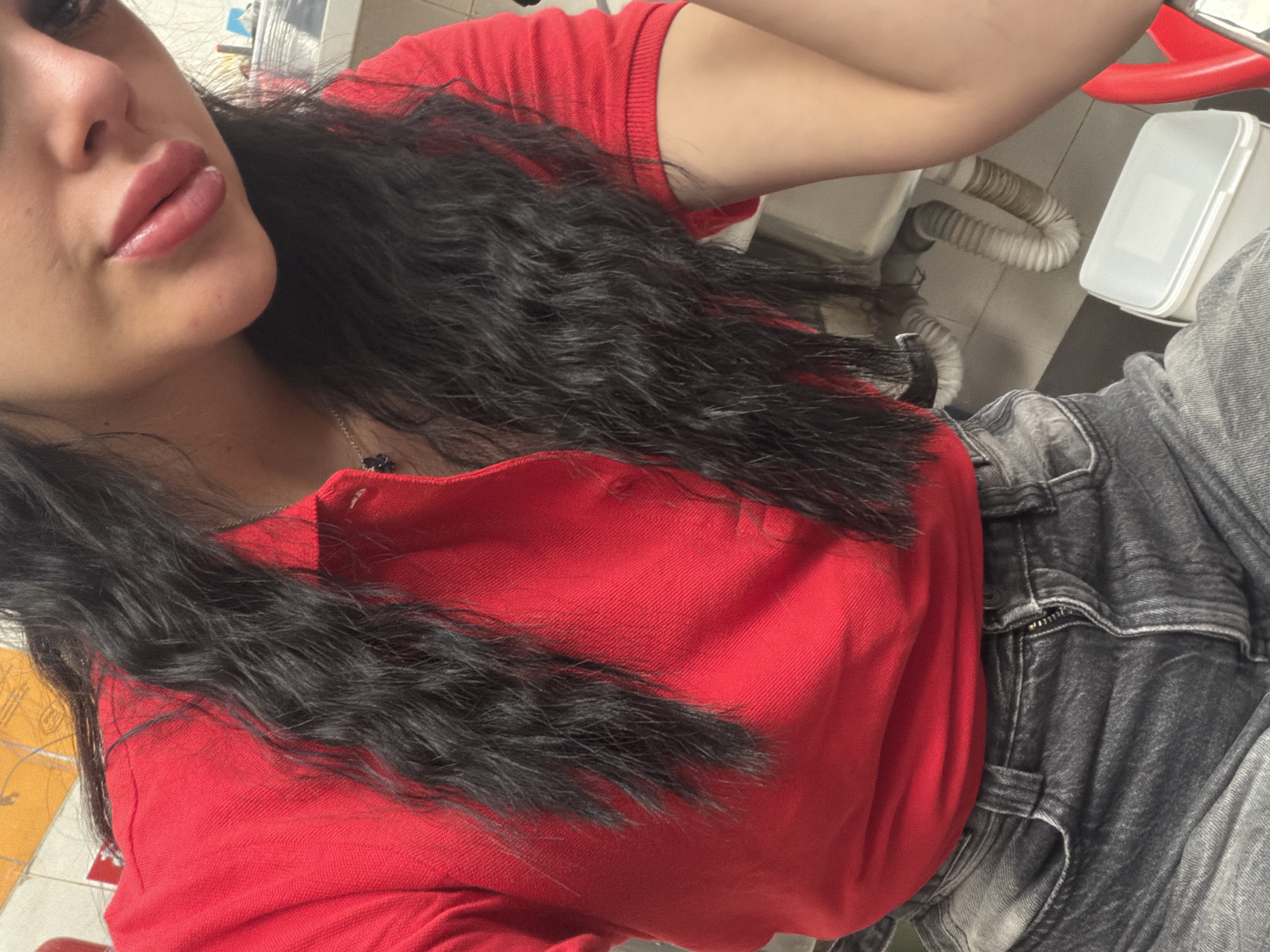 The image shows a close-up of a person wearing a red polo shirt and dark gray jeans. The person has long, dark, wavy hair and is wearing lipstick. The background is blurred but appears to be an indoor setting with plumbing fixtures and a white plastic container. The image is not specific enough to determine a location in Plovdiv, Bulgaria.