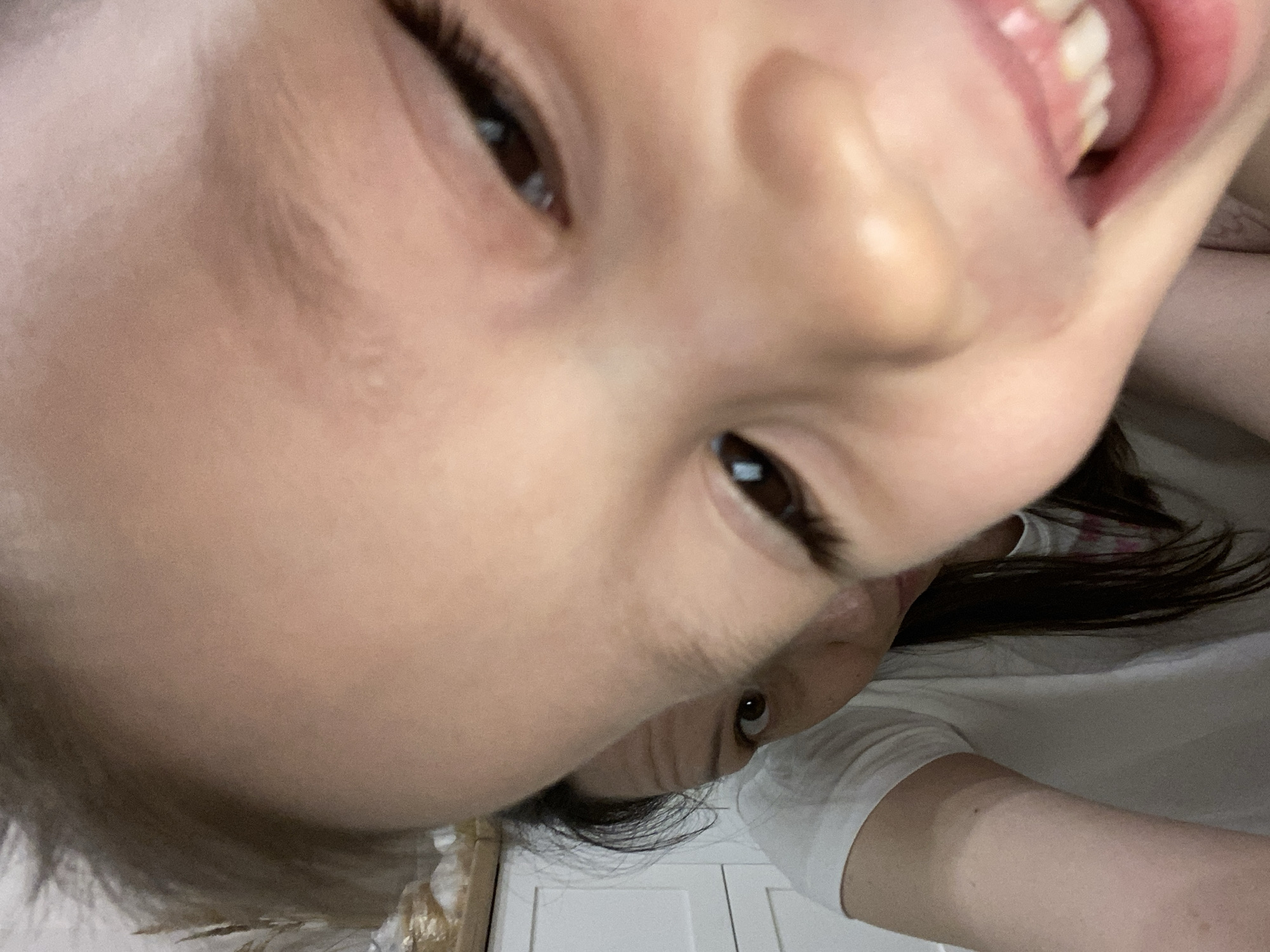 A close-up, downward-angled photograph shows a child's face with light skin, brown eyes, dark eyelashes, and an open mouth revealing teeth. Parts of an adult's face are also visible, including an eye and some hair. The adult is wearing a white t-shirt. A white door with panels is visible in the background. There is no evidence to confirm this photograph was taken in Chaikovsky, Russia.