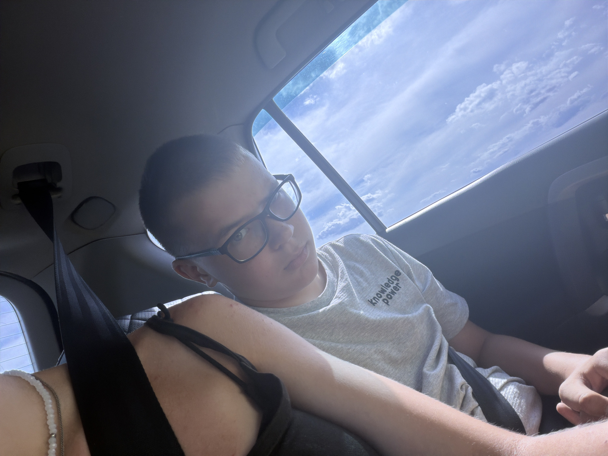 The image is a selfie taken from inside a car, likely in Novoe Teneevo, Russia. A young person, wearing glasses and a grey t-shirt with the words "Knowledge Power" printed on it, is visible in the foreground. The person is looking directly at the camera with a neutral expression. The car's interior is dimly lit, with a black seatbelt prominently displayed across the person's chest. Outside the car window, a bright blue sky with scattered white clouds is visible, suggesting it is daytime and likely sunny. The angle of the shot suggests the person is sitting in the back seat of the vehicle.