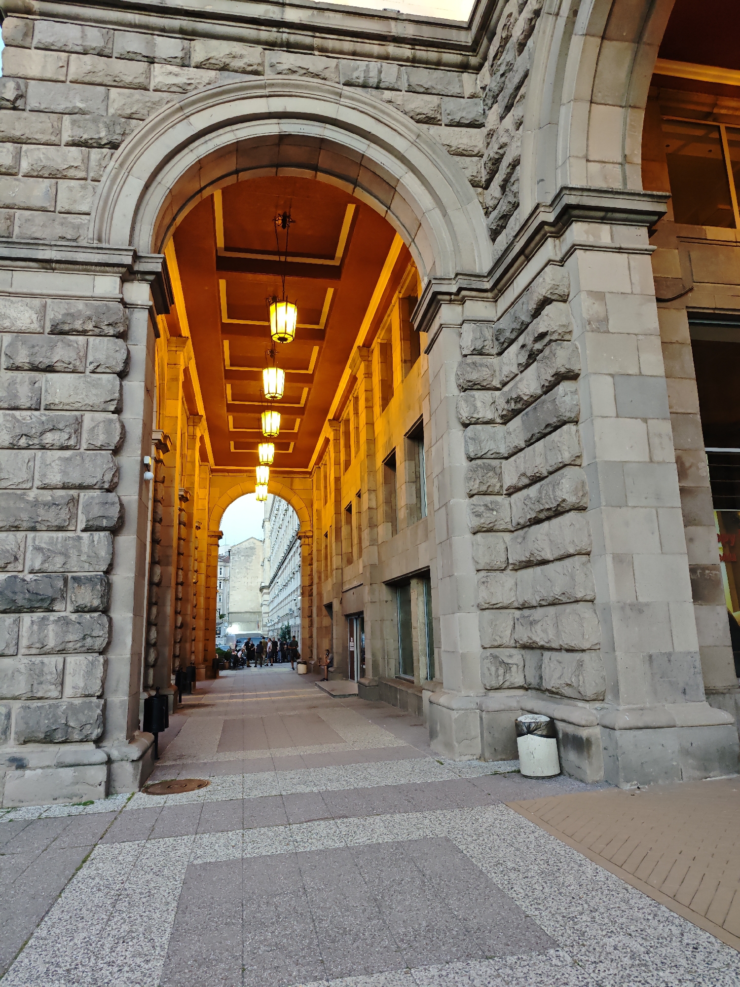 The image shows an arched pedestrian passage, likely in Sofia, Bulgaria, due to the architectural style commonly found in the city. The passage is constructed of large, rough-hewn stone blocks, giving it a monumental and somewhat imposing appearance. The interior of the passage is illuminated by a series of rectangular pendant lights with a warm, yellow glow, casting long shadows on the ceiling. The ceiling itself is dark brown with lighter brown rectangular accents. The floor of the passage is paved with interlocking tiles, featuring a geometric pattern of light gray and darker gray speckled sections.
On the left side of the passage, near the entrance, a black trash bin is visible. Further into the passage, on the right side, a light-colored, cylindrical trash receptacle stands. Beyond the immediate passage, a street scene unfolds, with a group of people gathered and conversing, some seated on benches. The background shows buildings with windows and what appears to be a building facade. The lighting suggests it is either early morning or late evening, with the artificial lights of the passage being prominent. The overall impression is one of an urban architectural feature, designed to provide shelter and a well-lit path through a larger structure.