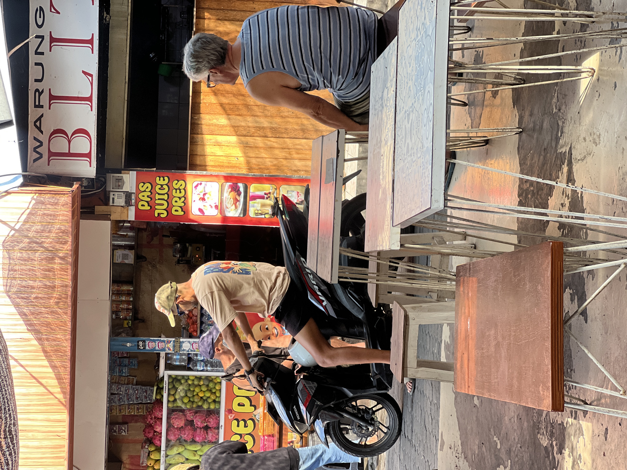 This image depicts a street scene in Kuta, Indonesia, characterized by a warung, which is a small shop or eatery, that appears to be selling fresh juices. The warung has a prominent sign that reads "WARUNG BLITZE," and a vertical sign advertising "PAS JUICE PRES." In front of the warung, there are several tables and chairs, some of which are empty and others are occupied.
A man wearing a light-colored t-shirt, shorts, sunglasses, and a cap is seated on a black Honda Vario scooter. He appears to be either waiting or about to depart. Behind him, another person wearing a blue cap is partially visible. To the right of the scooter, an older man with gray hair and glasses is seated at one of the tables, facing away from the camera. He is wearing a blue and white striped tank top and shorts.
The environment suggests a warm, outdoor setting, with sunlight casting shadows on the ground. The warung itself displays fresh fruits, including dragon fruit and oranges, suggesting the ingredients for the juices. The ground appears to be paved with stone or concrete and shows signs of moisture or staining. Several chairs and tables are arranged in front of the warung, indicating an outdoor seating area for customers. The overall atmosphere is that of a bustling street market or food stall area.