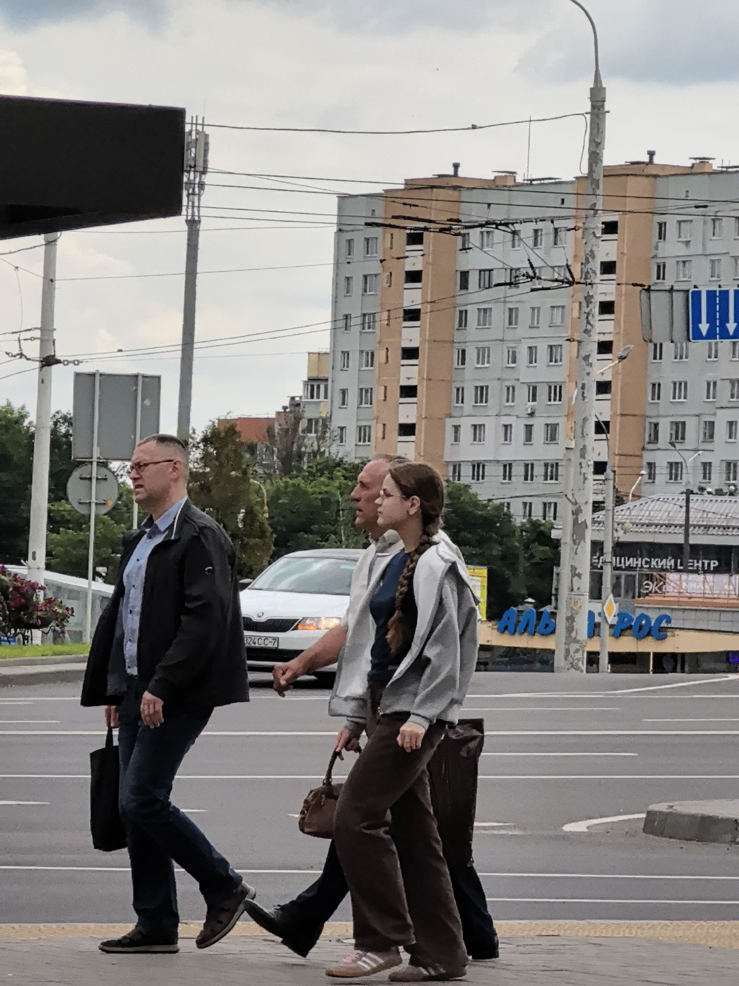 The image shows three people crossing a street in an urban setting. The people are walking from left to right across the frame. The person on the far left is a man wearing glasses, a blue collared shirt, and a dark jacket. He is carrying a black bag in his left hand. In the center is a younger person, possibly a woman, wearing a gray hooded jacket over a dark shirt and brown pants. She is carrying a brown handbag. To the right of this person is another individual, partially visible, wearing a dark jacket and blue jeans.
The background features a multi-story building with a light-colored facade and orange accents. Overhead, there are power lines and a lamppost. A white car is visible on the street, and a traffic sign with two blue arrows pointing downwards is present. The sky is overcast, suggesting a cloudy day. The scene appears to be a typical urban street crossing.
There is visible text on signs in the background. One sign, partially obscured, appears to read "МЕДИЦИНСКИЙ ЦЕНТР" in Cyrillic. Below this, another sign displays large blue letters that seem to read "АЛЬФА-ГРОС". The license plate on the white car is partially visible and reads "324 CC-7". The time of day is difficult to ascertain precisely, but the overcast sky suggests it is likely daytime, possibly afternoon. The overall mood is neutral, depicting everyday life.