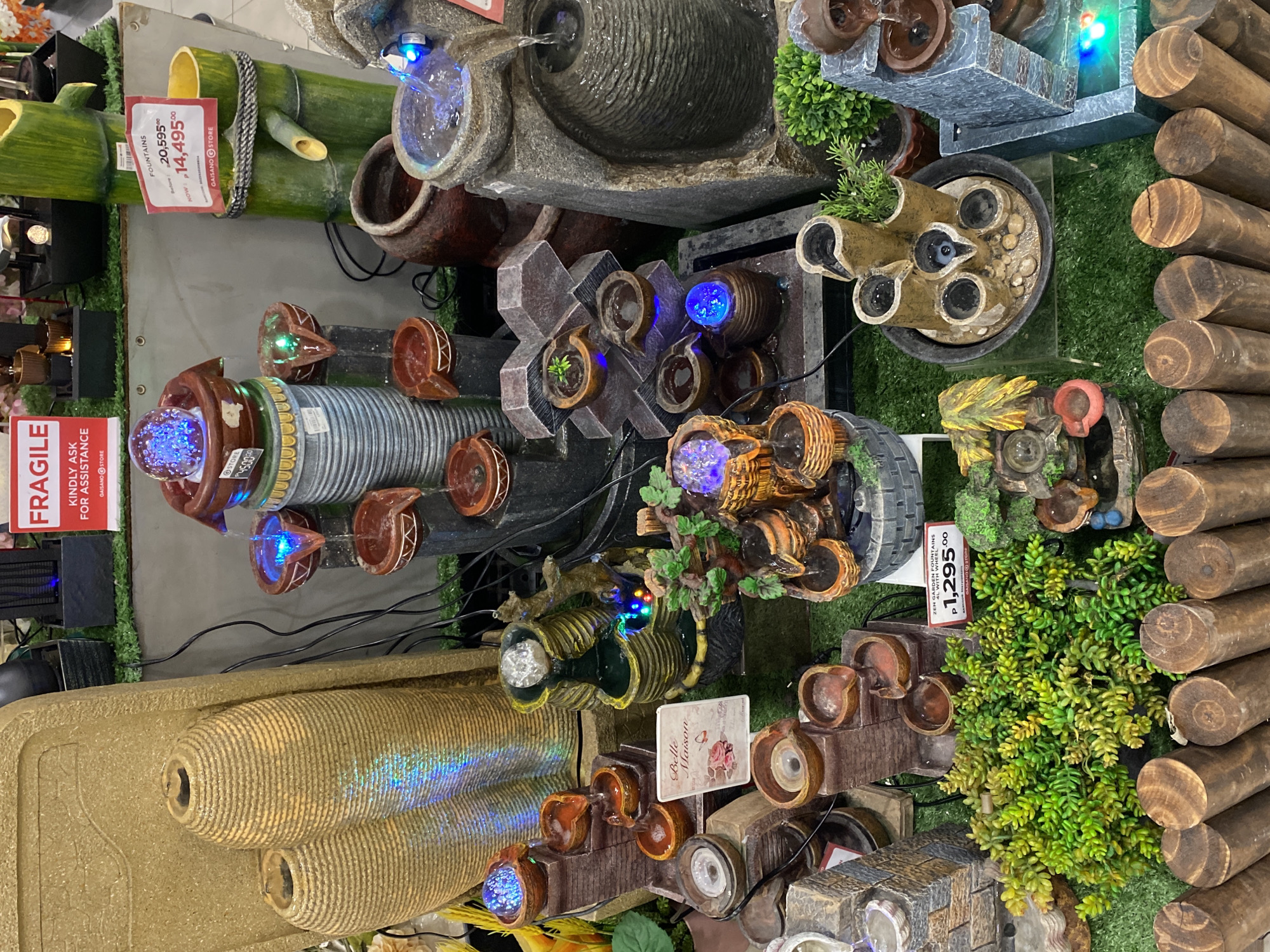 The image presents an indoor retail display of multiple decorative water fountains, likely situated within a Gaisano store in Davao City, Philippines, as indicated by "GAISANO STORE" branding on price tags and the use of Philippine Peso (P) currency. The display occupies a dedicated section, with the floor covered in artificial green turf and a low, wooden log-style fence bordering the immediate foreground.
Numerous water fountains of varying designs, sizes, and materials are showcased. These include multi-tiered structures resembling stacked stone, terracotta pots, bamboo chutes, and urns. Many of the fountains are operational, featuring circulating water illuminated by integrated LED lights that emit blue, green, and multi-colored glows. Several fountains incorporate transparent or clear glass spheres that also glow with internal lighting.
Prominent signage includes a red and white banner at the top reading "FRAGILE KINDLY ASK FOR ASSISTANCE." Various price tags are visible, such as a red tag for "FOUNTAINS" indicating a markdown from "P 20,595.00" to "P 14,495.00." Another red tag identifies "ZEN GARDEN FOUNTAINS 4L WITH WHEEL" priced at "P 1,295.00." A smaller white sign with a rose image reads "Belle Maison."
Decorative elements within the display include bundles of green artificial foliage, particularly succulent-like plants, placed around the fountain bases. On the far left, two tall, ribbed, tan-colored decorative vases are positioned. Black electrical cords connecting to the fountains are visible throughout the display area. No individuals are present within the captured scene.