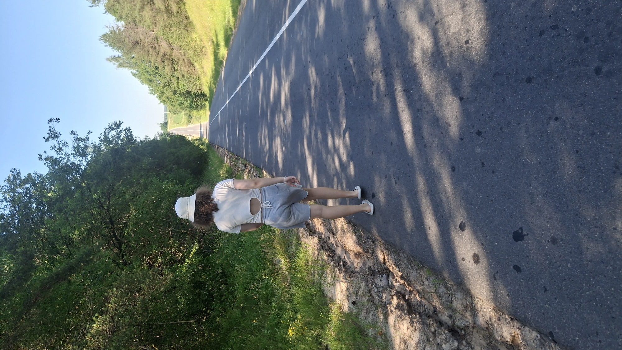 A person walks away from the camera down a paved road in a rural area. The person is wearing a light-colored bucket hat, a short-sleeved t-shirt with a cutout design on the back, and grey shorts. They are also wearing white flip-flops. The road has a white line down the center and a white line on the right edge. The road is flanked by trees and grass. The sky is clear and blue. The lighting suggests it is daytime, possibly late afternoon, as indicated by the shadows on the road. The surrounding environment appears to be natural, with no visible buildings or infrastructure other than the road itself. The location is identified as Дзямідавіцкі сельскі Савет, Belarus.