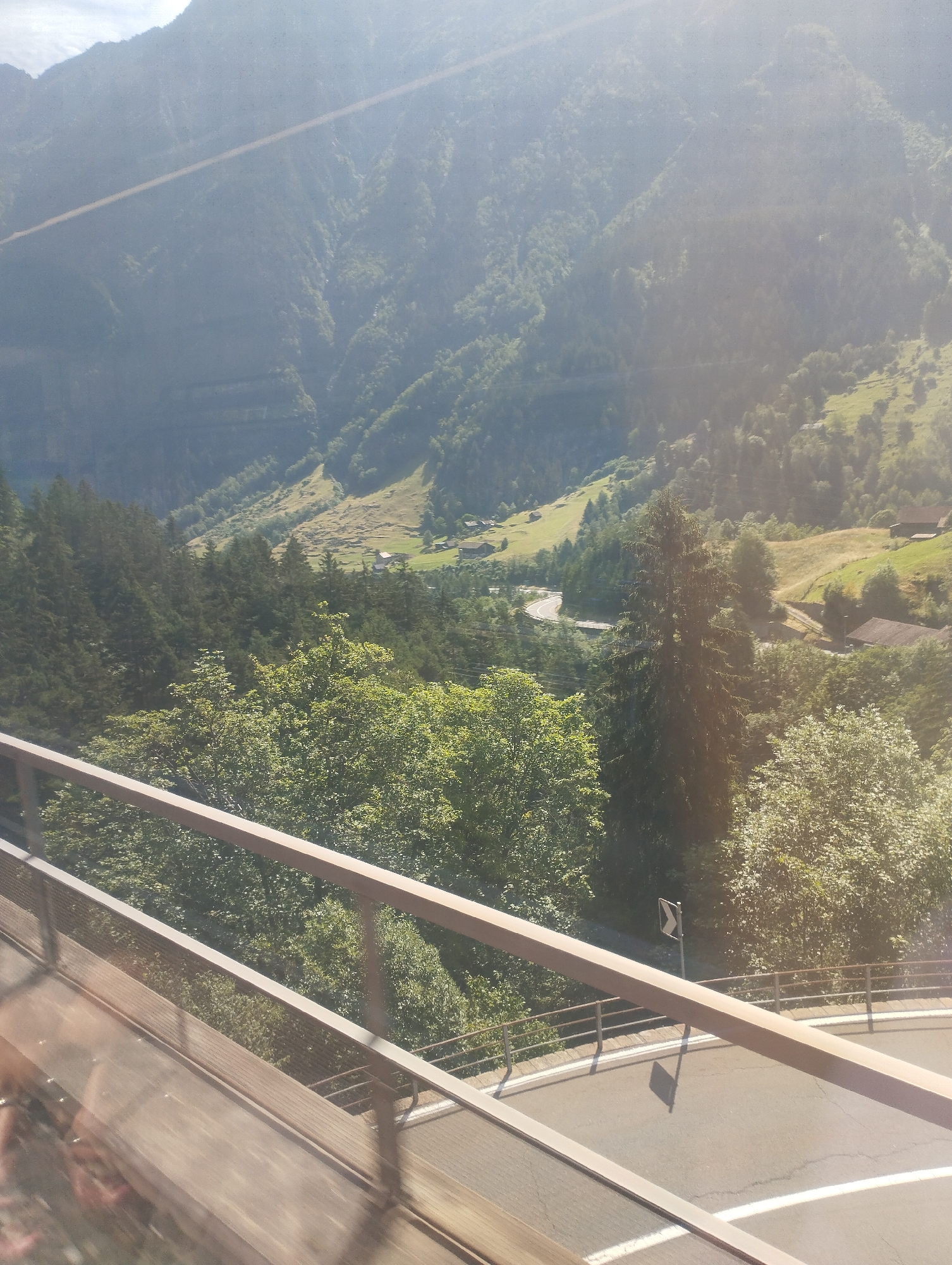 The image depicts a scenic mountain landscape in Wassen, Switzerland. The scene is viewed from an elevated position, possibly a balcony or a train. In the foreground, a wooden railing and a metal barrier are visible, suggesting an observation point or walkway. Below the railing, a paved road with white markings curves through the scene, indicating it's a mountain pass or a road winding through the valley.
The midground is dominated by lush green trees, a dense forest covering the slopes of the mountains. Among the trees, hints of a river or stream can be seen, reflecting the sunlight. Further into the background, the mountains rise dramatically, their slopes covered in a mix of trees and grassy meadows. Sunlight streams in from the top left, casting a bright glow and creating lens flare, suggesting it's a sunny day, likely late morning or early afternoon due to the angle of the light.
Small houses with red roofs are scattered across the valley floor and on the lower slopes, hinting at a rural settlement. A road sign with a black arrow on a white background is visible near the winding road, indicating a sharp turn. The overall atmosphere is one of natural beauty and tranquility, capturing the essence of the Swiss Alps. There is no visible text in the image other than the road sign.