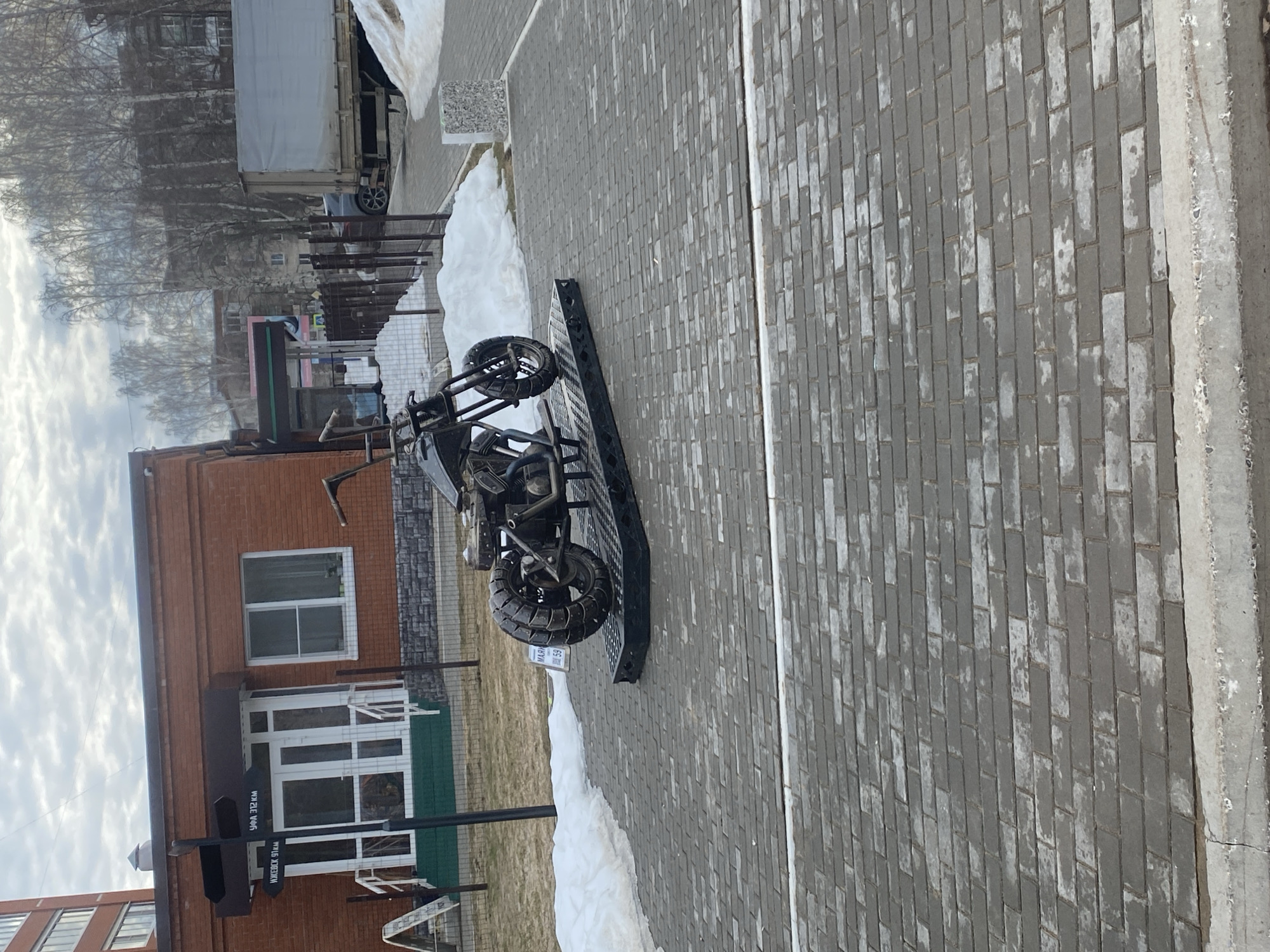 The image shows a black trike-style motorcycle parked on a paved brick sidewalk in Chaikovsky, Russia. The motorcycle is resting on a metal ramp. Behind the motorcycle is a brick building with white-framed windows and a black sign with white Cyrillic text. Patches of snow are visible along the edge of the sidewalk and near the building. Further back, trees and a truck can be seen. The sky is overcast.