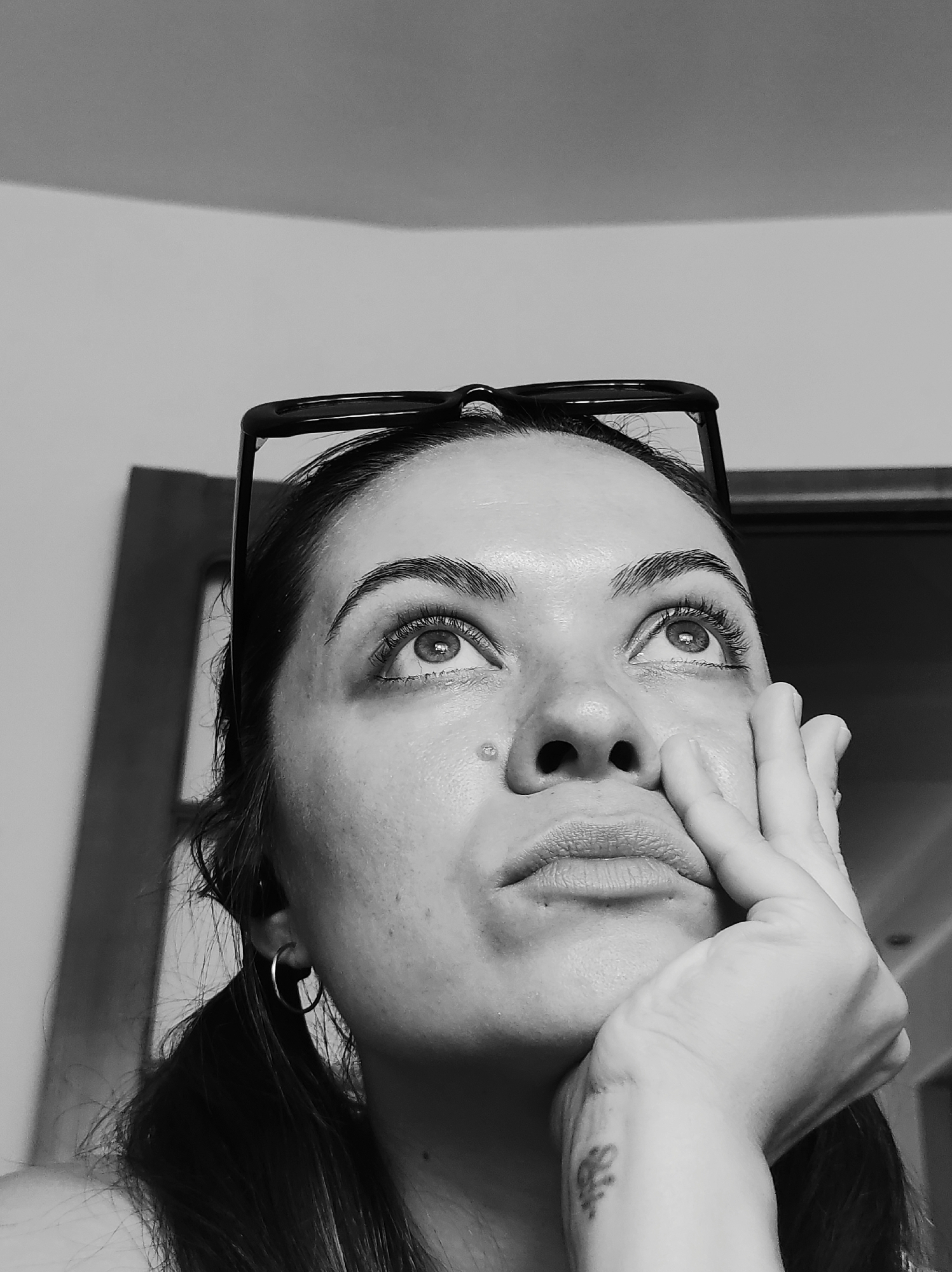 The image is a black and white, close-up, low-angle shot of a woman's face from the shoulders up. She has her head tilted back and is looking upwards. Her right hand is resting on her chin and cheek, with her fingers extended. She is wearing a pair of glasses perched on top of her head. Her hair is pulled back into two low ponytails, and she is wearing a hoop earring in her left ear. There is a small mole on her cheek and a faint tattoo on her left wrist. The background is blurry but appears to be an indoor setting with a wall and a doorframe. There is no discernible information about the location being in Chaikovsky, Russia.