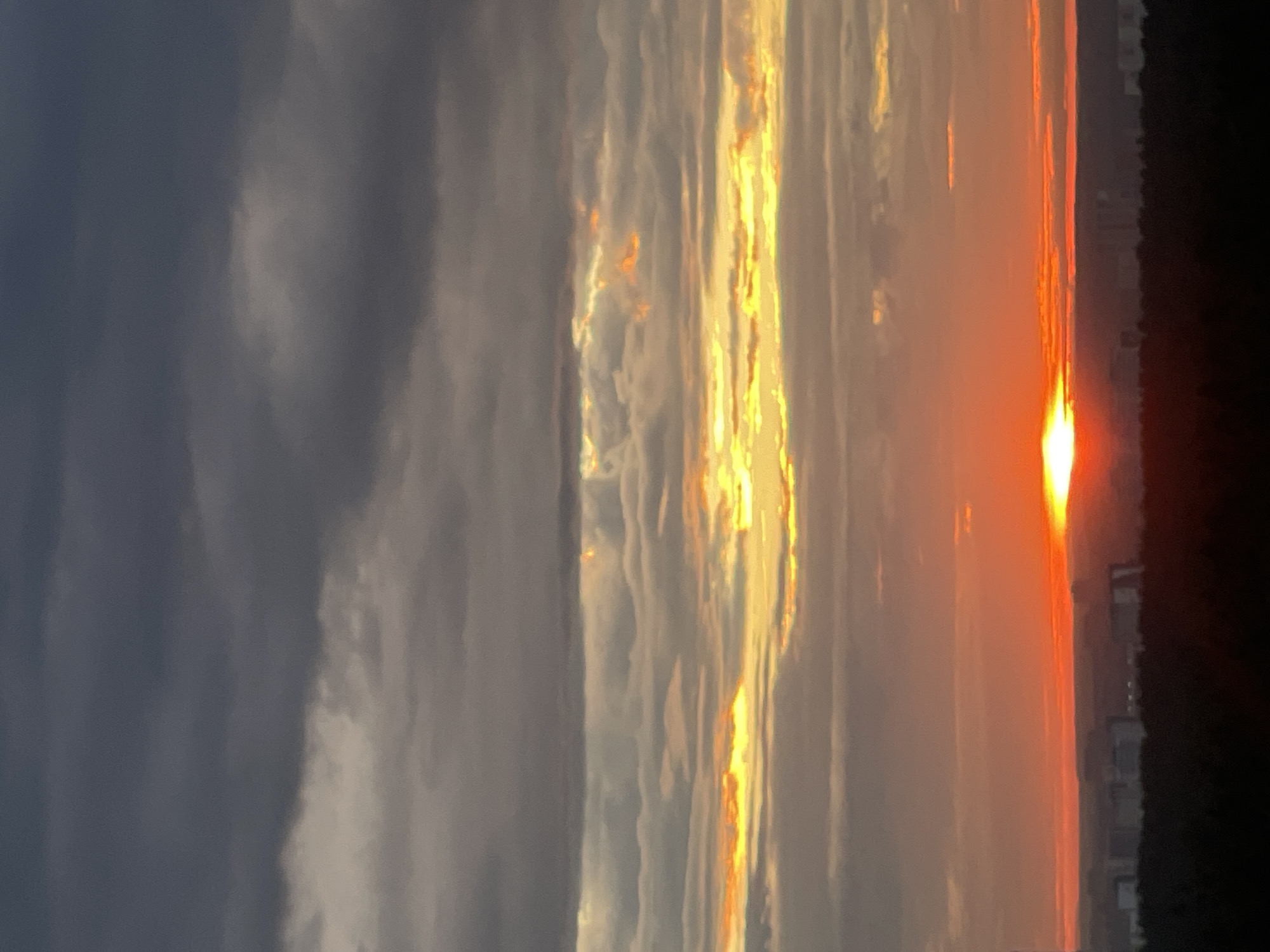The image shows a cloudy sunset over the city of Perm, Russia. The sky is filled with layers of dark gray and lighter gray clouds, with streaks of orange and yellow light breaking through near the horizon. The sun is partially obscured by clouds, casting a warm glow over the scene. Below the sky, a dark silhouetted treeline stretches across the bottom of the frame, with the faint outlines of buildings visible beyond it, suggesting the city's sprawl. The overall mood is somber yet beautiful, characteristic of a dramatic sunset. There are no people or specific actions depicted in the image.