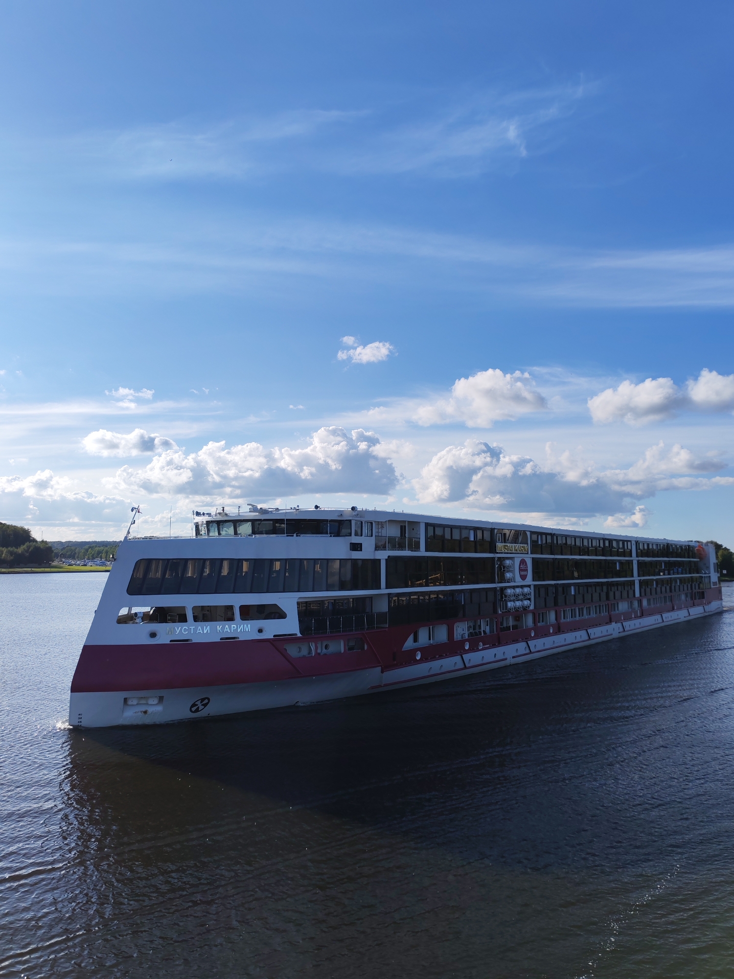 A large, white and red passenger ship named "МУСТАЙ КАРИМ" (Mustai Karim) is seen from a slightly elevated angle, traveling on a body of water. The ship has multiple decks with numerous windows and appears to be in motion, creating a wake. The sky above is bright blue with scattered white clouds. In the distance, the shoreline is visible with trees and some structures, suggesting a semi-urban or rural environment. The water is dark and reflects the sky and the ship. The name of the ship is prominently displayed on its hull. The ship is the primary subject, dominating the center and right portion of the frame. The water occupies the lower part of the image, and the sky fills the upper portion. The overall scene depicts a serene day of river or lake travel.