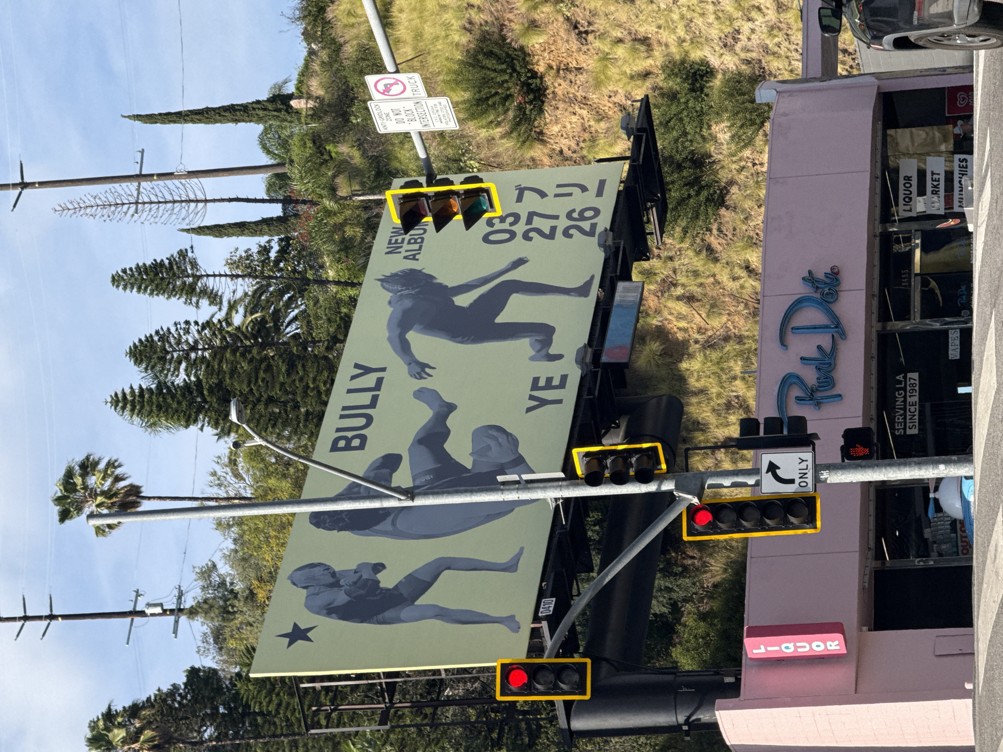 Here is a detailed description of the image:
**Scene:** The image is taken in West Hollywood, California, USA, focusing on an intersection where a large billboard dominates the view. The billboard advertises a new album, "BULLY YE," with the release date of March 26, 2024, featuring stylized silhouettes of figures engaged in what appear to be wrestling or martial arts maneuvers.
**Objects:**
- **Billboard:** Large and centrally located, featuring the album advertisement.
- **Traffic Lights:** Multiple sets of traffic lights with red and partially visible green lights. They are attached to metal posts and are highlighted.
- **Signs:** There are several signs, including a "Turn Only" sign and an anti-gridlock zone sign.
- **Building:** A pink building labeled "Puck Daugh" and "LIQUOR MARKET" with signs also indicating "SERVING LA SINCE 1987" and "MUNCHIES," along with "VAPES."
- **Vegetation:** Trees of various types are seen in the background and to the sides of the billboard, suggesting a residential or semi-urban setting.
- **Other Objects:** Utility poles with electrical wires, sections of a car, and parts of the street are visible.
**Environment:** The scene is set during daylight under a partly cloudy sky. The general atmosphere is urban, with a mix of commercial and natural elements.
**Interactions:** The traffic lights suggest regulated movement, and the presence of the billboard indicates commercial advertising. The lack of visible people in the immediate foreground implies that this is a moment frozen in time, capturing the intersection without pedestrian or vehicular activity.
