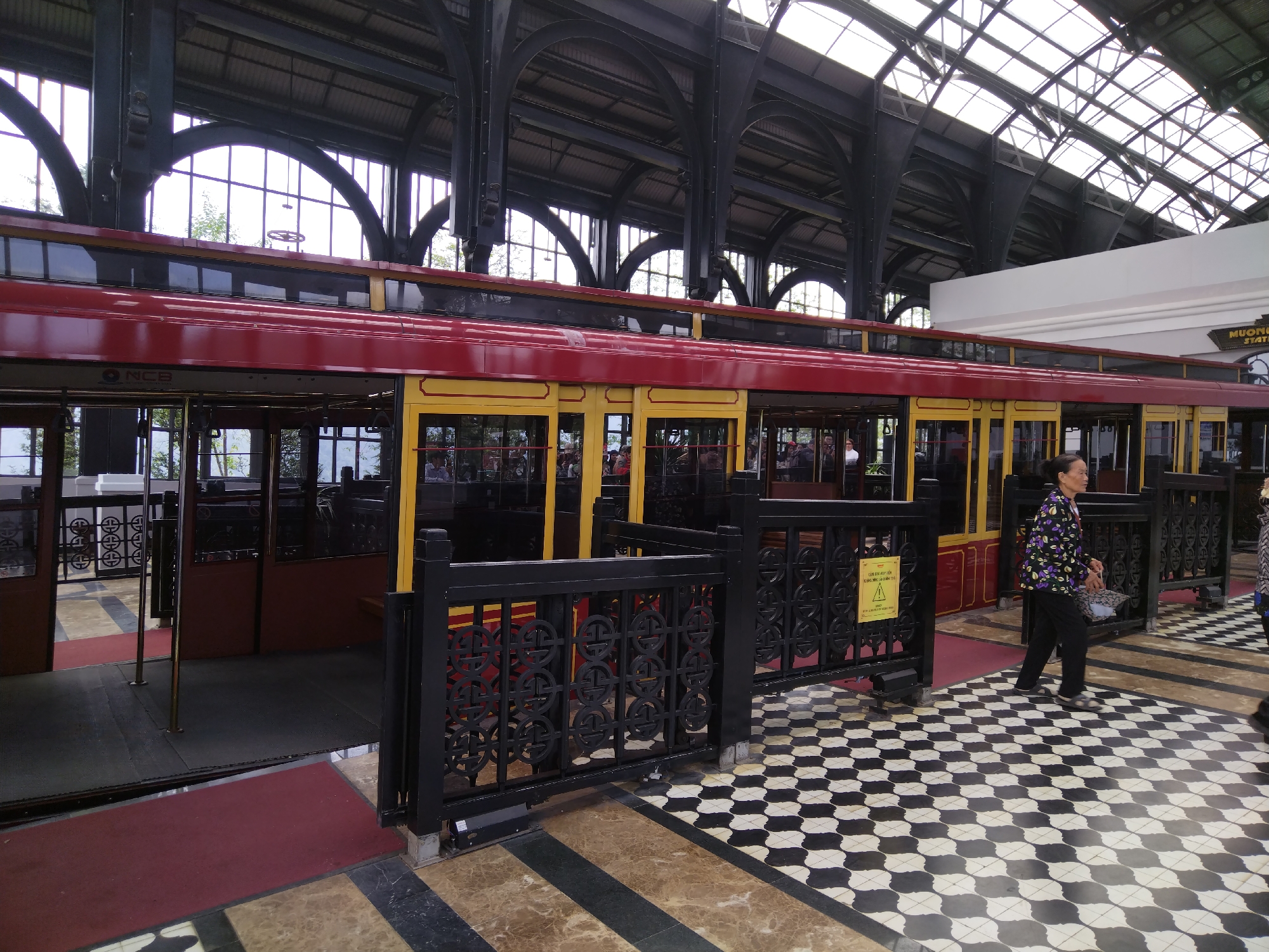 The image shows a railway platform with a red and yellow train car, an older woman in a patterned jacket, and a black and white checkered floor. The train car is partially open, and passengers are visible inside. A black fence separates the platform area. The architecture of the station features arched steel beams and a skylight roof. The location is likely a tourist attraction or a historic railway station.