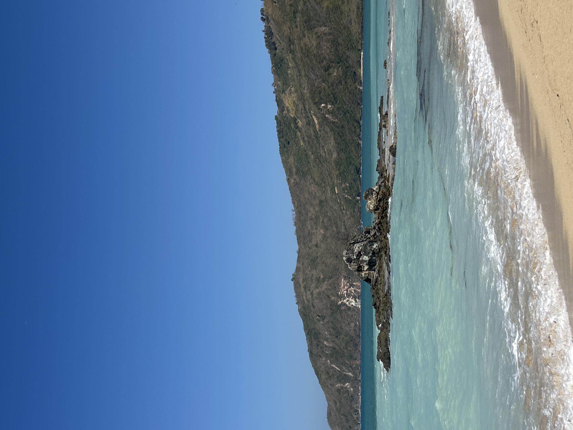 The image depicts a serene coastal landscape. In the foreground, a sandy beach transitions into shallow, turquoise water with gentle waves lapping at the shore. Further out, a cluster of dark rocks emerges from the sea, some partially submerged. Beyond the rocky outcrop, the water deepens to a richer blue. In the background, a verdant, hilly landscape dominates the horizon under a clear, bright blue sky. The hillside appears dry and sparsely vegetated in some areas, with some patches of lighter-colored rock or soil visible. There is no indication of the city of Kute, Indonesia, or any specific landmarks within the image. The scene is devoid of any people or human activity.