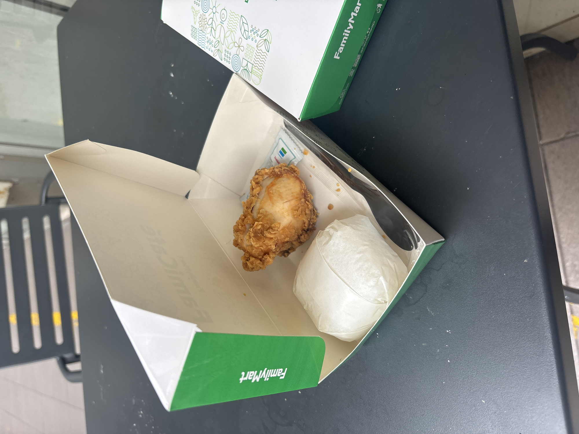 A close-up, eye-level shot shows a FamilyMart bento box with a fried chicken piece and a rice ball wrapped in white paper, placed on a dark gray textured table. A small packet of sauce is visible next to the chicken. To the right, another partial FamilyMart box with a green and white design is visible, suggesting a meal bought from the convenience store. The background shows a dark gray chair with yellow stripes and a portion of a tiled floor. The image appears to be taken outdoors, possibly at a FamilyMart branch in North Jakarta, Indonesia, given the context of the brand and typical urban outdoor seating. The lighting suggests it is daytime.