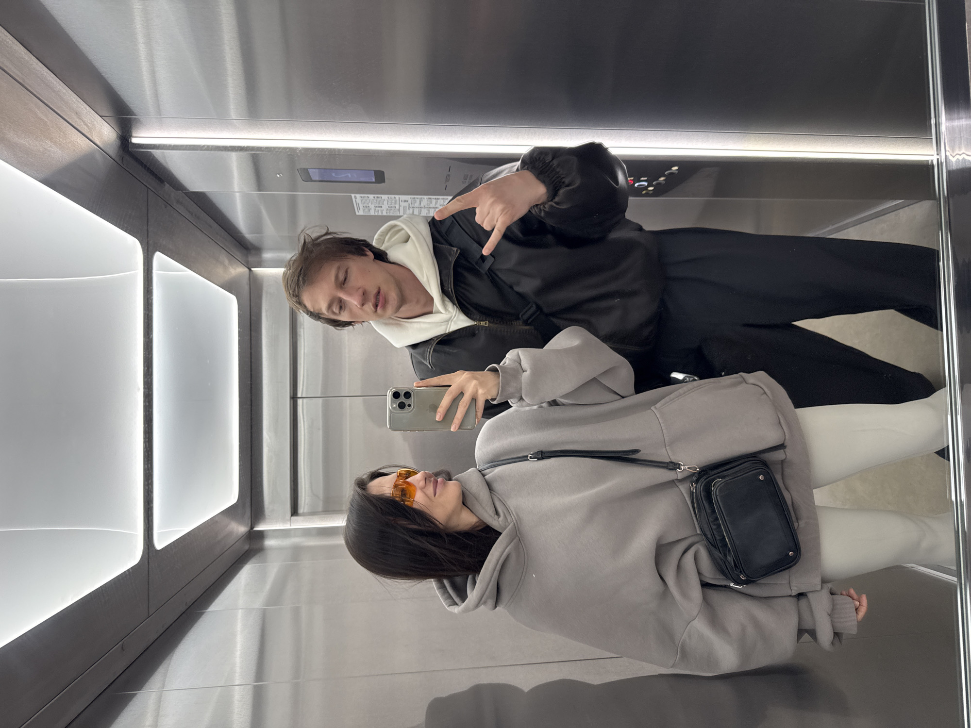 Two individuals are inside an elevator with stainless steel walls and ceilings. A man, wearing a black jacket over a white hoodie and black pants, is posing with a hand gesture. A woman, wearing a gray hoodie, white pants, and orange-tinted glasses, is holding a cell phone, presumably taking the photograph of themselves. A black shoulder bag hangs from her hoodie. Elevator buttons are visible in the background, along with the floor indicator and other elevator fixtures. The location, Murino, Russia, is not deducible from the image.