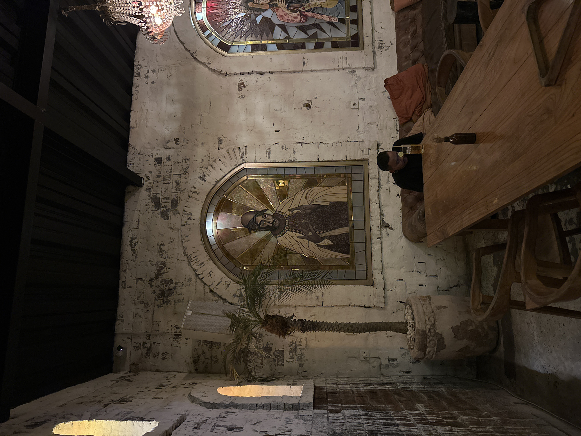The image captures an indoor space with a distinctive, rustic ambiance. The walls are made of distressed, white-washed brick or stone, showing considerable texture and wear. The ceiling is high, with dark, possibly slatted panels visible on the far left.
Two prominent arched stained glass windows adorn the wall. The upper window depicts a figure playing a stringed instrument, rendered with a halo-like background. The lower window features a bearded figure with a similar radiant background, extending an open hand, stylized in a manner reminiscent of religious iconography but resembling cultural figures such as Tupac Shakur.
An ornate chandelier, possibly adorned with crystals or beads, hangs from the ceiling in the upper left, emitting a warm glow. Below the lower stained glass window, a slender palm tree stands in a weathered, decorative planter on the floor.
On the right side of the frame, a person with dark hair and an ear gauge, wearing a dark long-sleeved shirt, is seated at a large, sturdy wooden table. They appear to be looking down. On the table in front of them is a glass containing a drink with ice and a lime wedge, alongside a dark glass bottle. Upholstered seating with a reddish-brown cushion is visible around the table.
The lighting is dim and warm, suggesting an evening or nighttime setting. The overall scene evokes a relaxed and atmospheric mood, characteristic of a unique bar, restaurant, or themed establishment. No visible text is present in the image.
