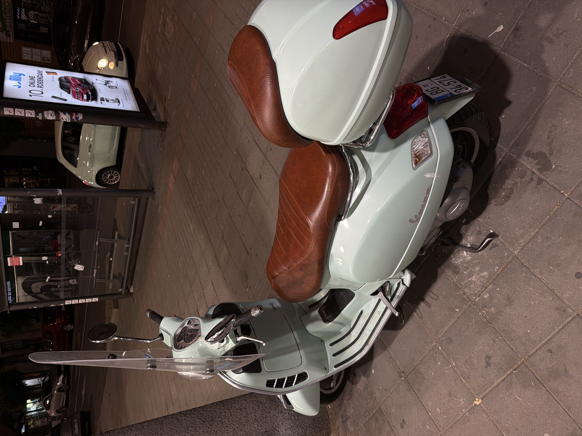 A mint green Vespa scooter with a brown leather seat and a rear storage compartment is parked on a tiled sidewalk at night in Vracar Urban Municipality, Serbia. The scooter is angled to the right, with its front facing slightly left. A tall, clear windscreen extends upwards from the handlebars. The background includes a bus stop shelter with benches, streetlights, and parked cars, suggesting an urban street scene. A poster advertising "Lilly online rođendan" is visible on a pole near the bus stop. The license plate of the scooter reads "BG 34-574".
