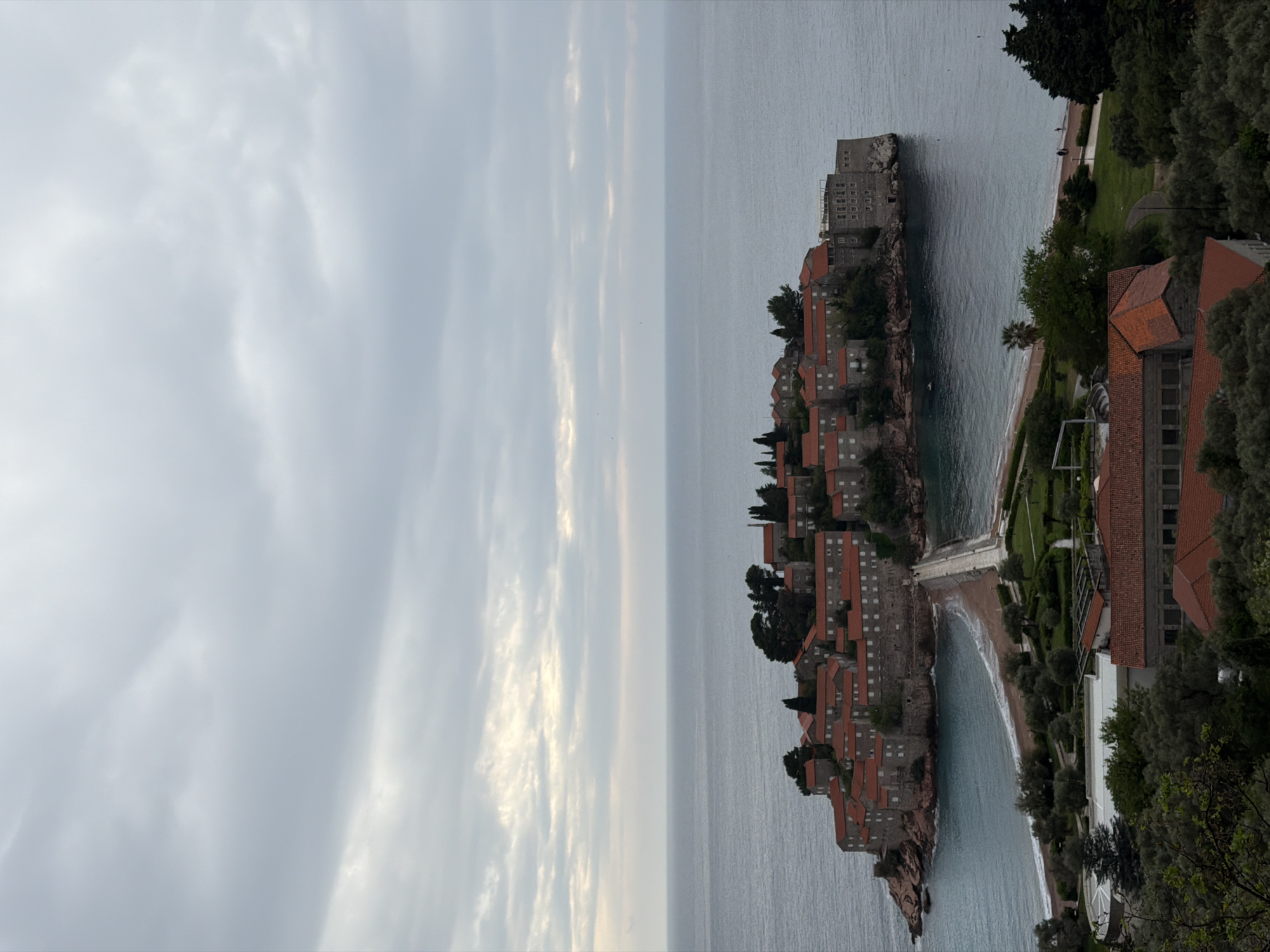The image displays an elevated view of the iconic islet of Sveti Stefan in Montenegro. The islet is densely covered with numerous historic stone buildings, each featuring traditional terracotta-tiled roofs. These structures are nestled together, extending into the calm, grey-blue waters of the Adriatic Sea.
A narrow, sandy causeway connects the islet to the mainland. On the mainland side, the landscape is characterized by lush green trees and manicured grounds. Several additional buildings are visible, including one with a red-tiled roof similar to those on the islet, and another with a lighter-colored facade. Two crescent-shaped beaches flank the causeway, meeting the gentle waves of the sea. The sea itself appears placid, stretching towards a hazy horizon.
The sky is uniformly overcast with soft, diffused clouds, suggesting a tranquil, cloudy day, possibly during the morning or late afternoon, as there are no harsh shadows. The overall atmosphere conveys serene beauty and historical charm. A few very small, indistinct figures might be present on the mainland beach or paths, but no specific activity is clearly discernible. There is no visible text within the image. The unique architectural style and the distinctive island-causeway formation are clear location cues for Sveti Stefan.