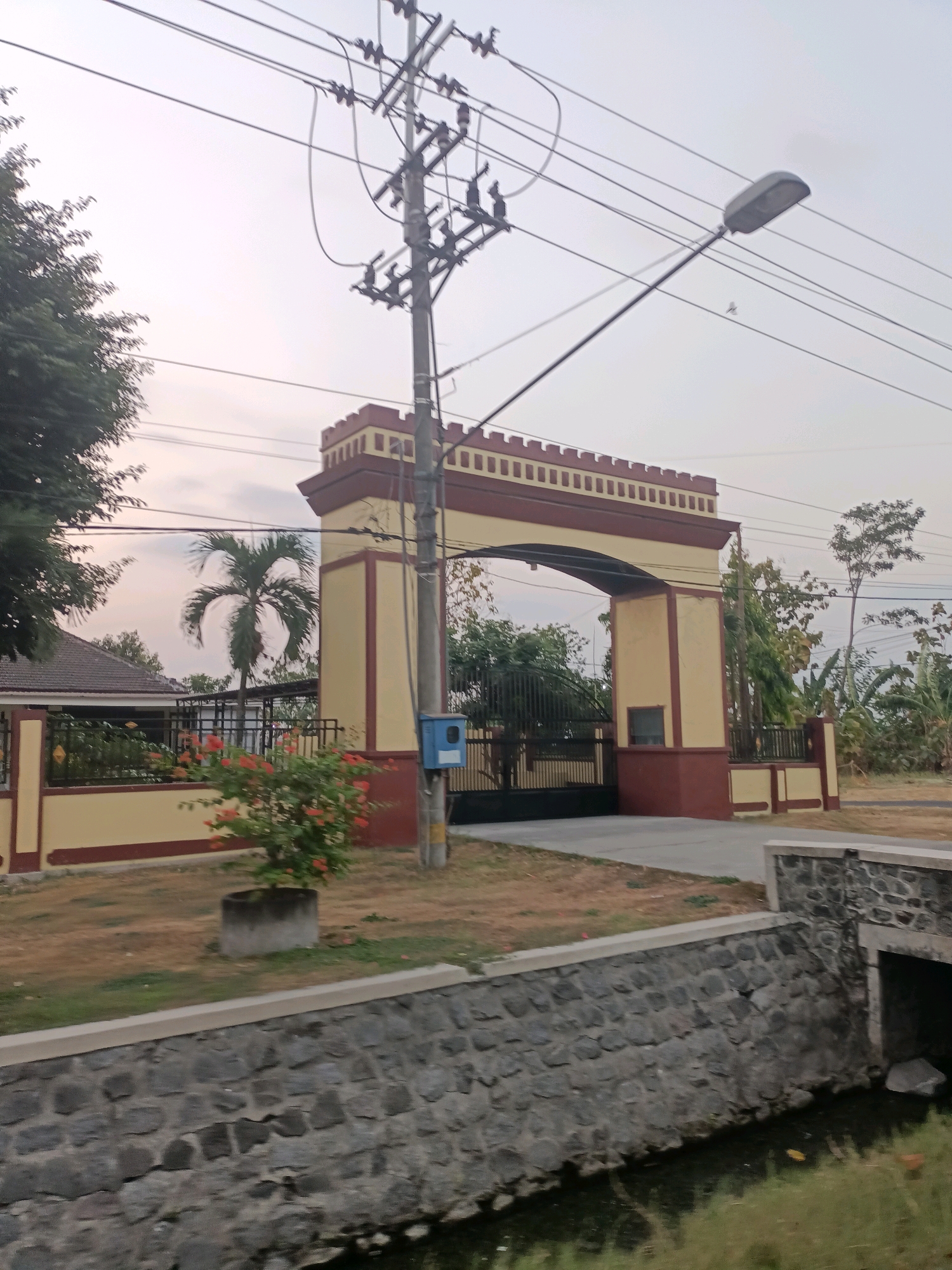 The image captures a grand archway, likely an entrance to a facility, located in Badas, Indonesia. The archway is painted in shades of cream and maroon, with crenellated battlements along the top, giving it a castle-like appearance. It is flanked by pillars and a wall. A black metal gate is visible through the arch. To the left of the arch, a palm tree stands tall, and a residential building with a red roof is partially visible, bordered by a black fence. In front of the wall, a small plant with orange flowers in a concrete planter adds a touch of color. A blue electrical box is mounted on a utility pole that also supports numerous overhead wires and a street light. A stone wall runs along the foreground, bordering a small canal or drain. Trees and foliage are scattered throughout the background, suggesting a semi-urban or suburban environment. The sky is overcast, indicating diffused daylight. There are no people visible in the scene.