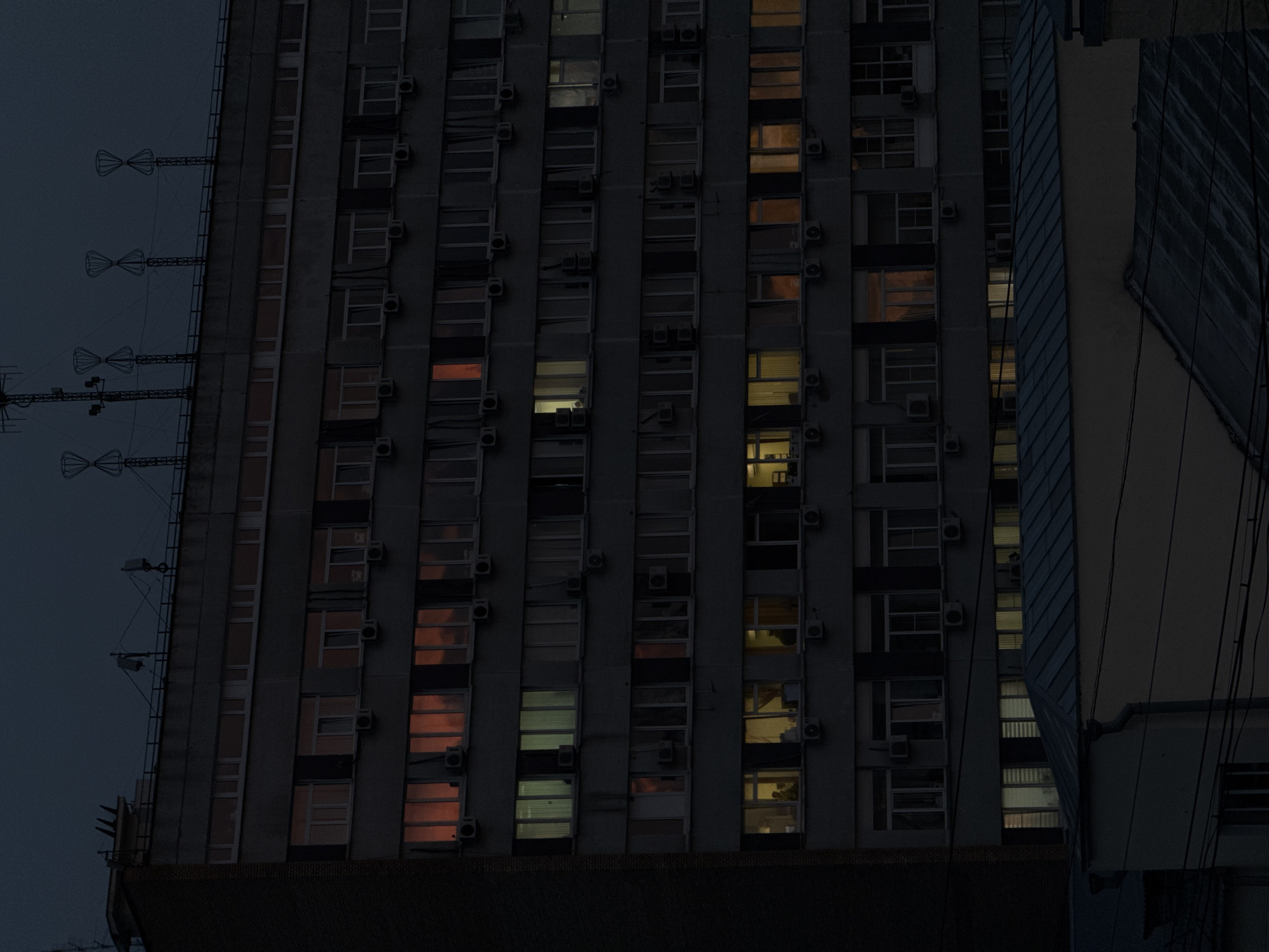 The image depicts a tall building in Moscow, Russia, at twilight. The building is characterized by numerous windows, some of which are illuminated from within, casting a warm glow. Air conditioning units are attached to the exterior of the building. A series of antenna-like structures is visible on the left side of the building. A second building is partly seen on the right, separated from the main building by a few thin, dark wires. The overall lighting suggests it is either early evening or late dawn, with a dark sky providing the backdrop.