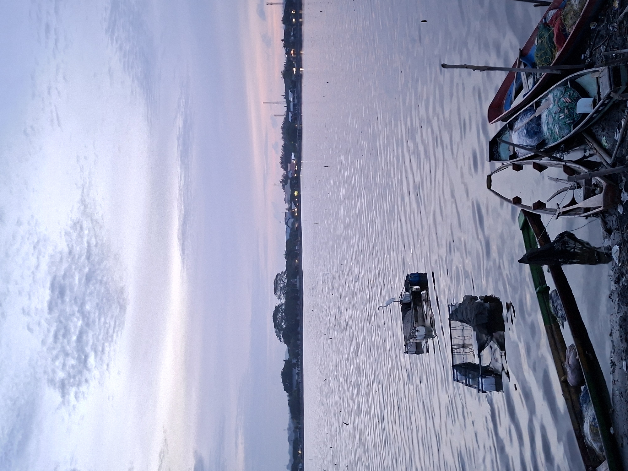 The image depicts a body of water, possibly a river or lake, with a shoreline in the distance featuring buildings and lights, suggesting a town or city. Several boats are visible, both near the shore and further out in the water. Some boats are small and appear to be moored near the shoreline, while others are more distant and might be in transit. The sky is overcast with muted colors, indicating either dawn or dusk. The water surface has small ripples, suggesting a light breeze. The overall scene has a calm, tranquil ambiance. The location is Lhokseumawe, Indonesia.