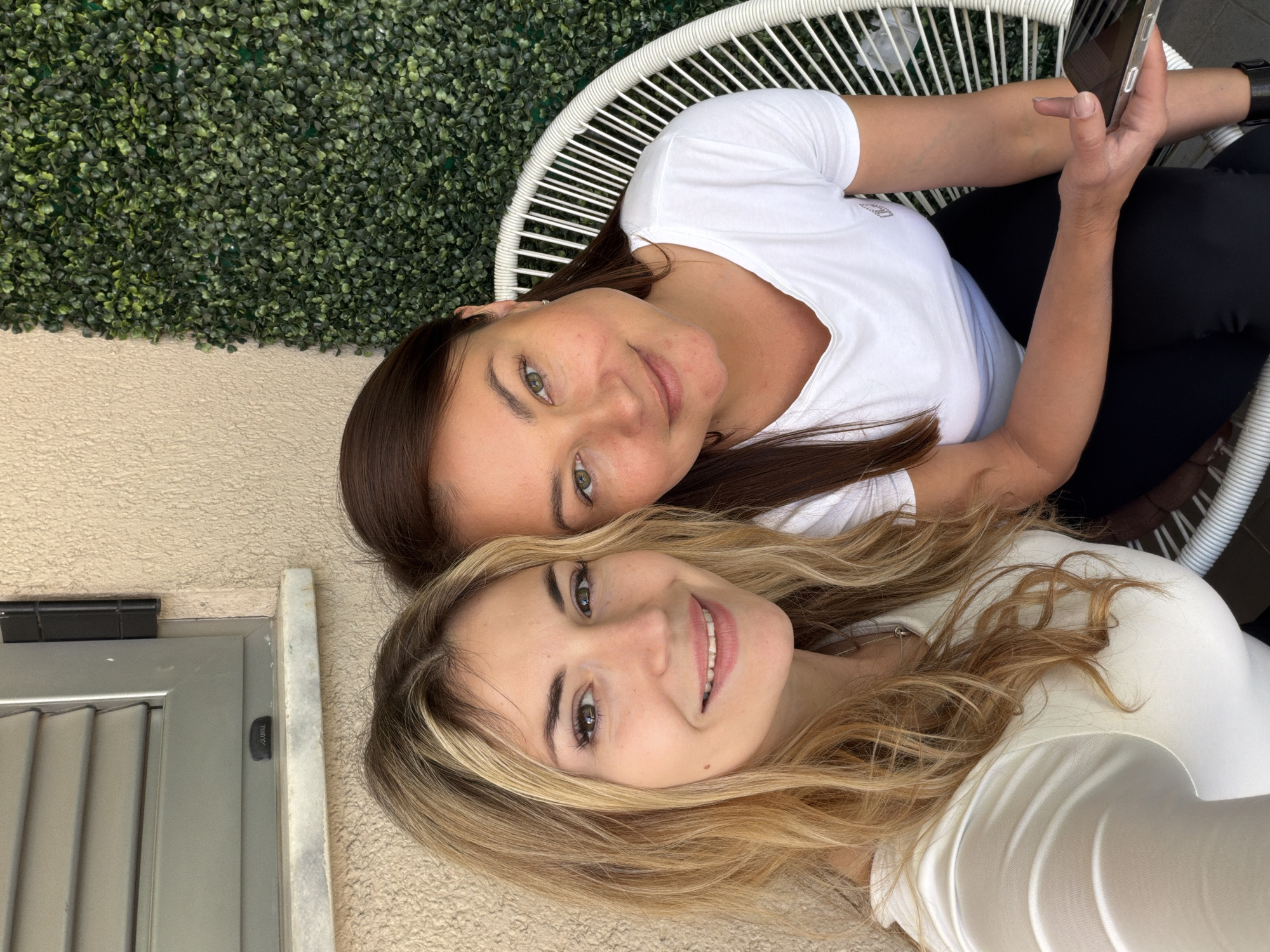 Two women are posing for a photo, likely a selfie, outdoors. One woman has long brown hair, green eyes, and is wearing a white t-shirt and black pants while seated in a white wicker chair. She holds a smartphone in her hand. The other woman has long blonde hair, light-colored eyes, and a white top. Part of a green wall is visible in the background, along with a beige-colored wall with a door. It is likely that the picture was taken in Budva, Montenegro.