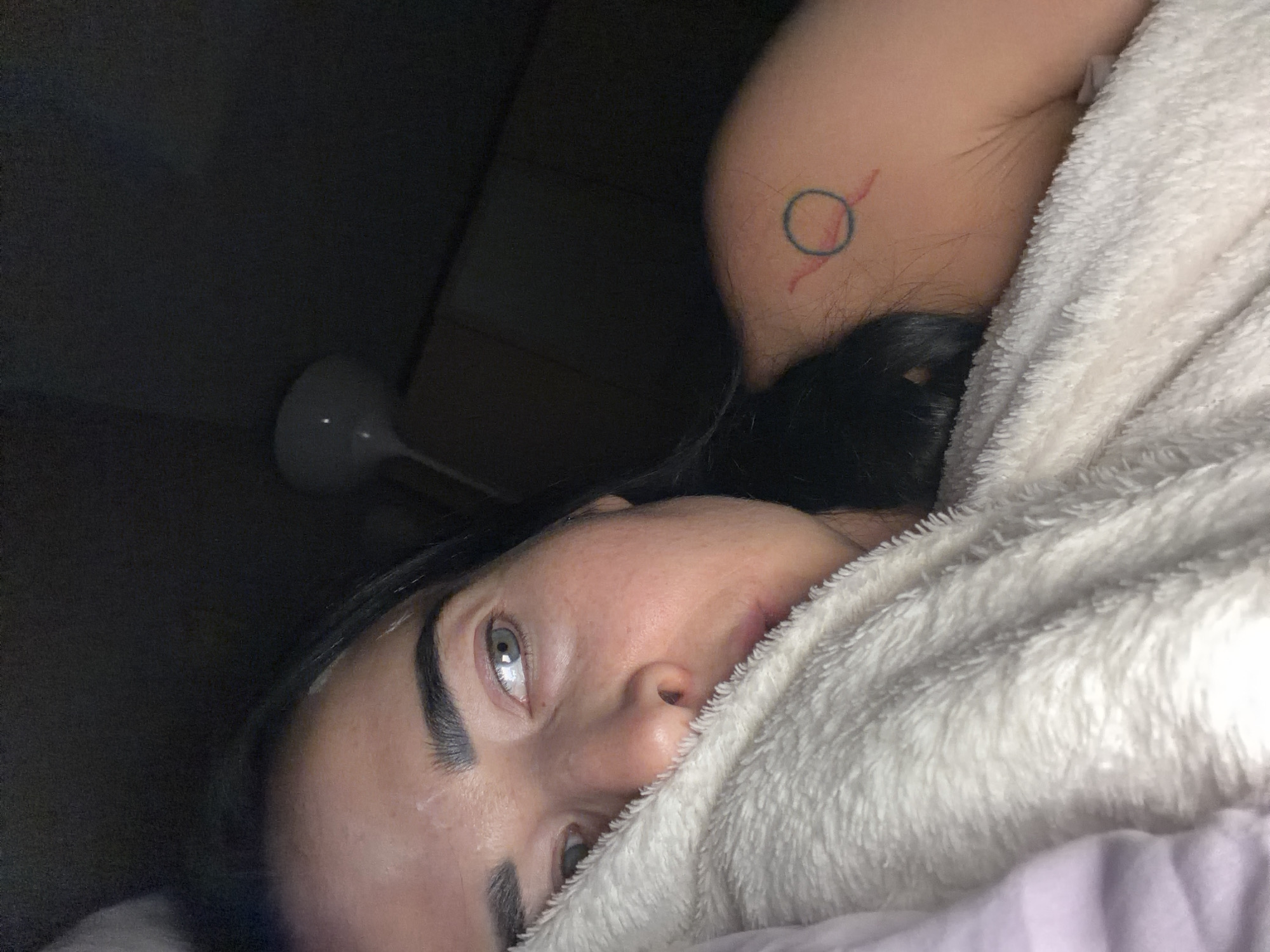 A close-up, dimly lit image shows a person lying down, partially covered by a fluffy white blanket. The person's face, primarily their left eye, nose, and dark eyebrows, is visible in the foreground, angled upwards. Their light-colored eye (appearing blue or grey) is open, reflecting a small light source. Dark hair is visible behind and around their head.
On the upper arm/shoulder area, partially obscured by the blanket, a small tattoo is visible. It consists of a blue circle with two red, stylized, slightly wavy or jagged lines extending outwards from it. The skin around the tattoo appears pale with some hair.
The background is very dark, suggesting a low-light environment, possibly nighttime or a room with minimal illumination. A portion of a light-colored, modern-style lamp with a rounded head is discernible in the dark upper left corner, contributing to the minimal light in the scene. The overall impression is one of a person resting indoors. No text is visible in the image.