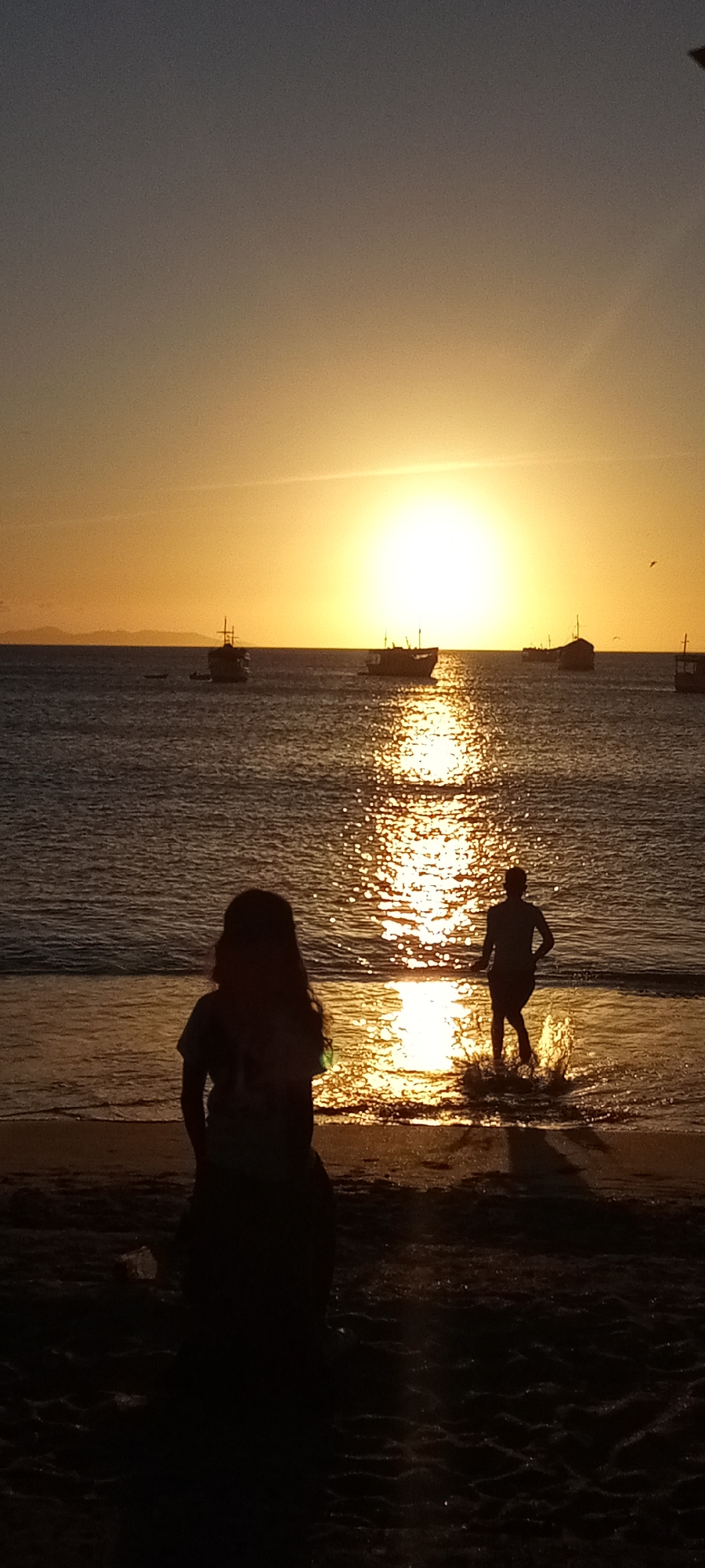 At a beach in an unknown location, the sun is setting over the ocean, casting a golden light. Several fishing boats are visible on the horizon, silhouetted against the bright sunlight. In the foreground, a woman stands on the sandy beach, looking out at the ocean. A man is running into the water, kicking up a splash.