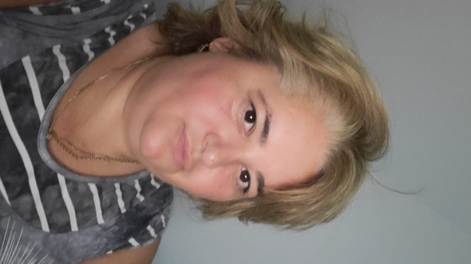 A medium shot portrait of a woman with shoulder-length blonde hair, her gaze directed towards the camera. She is wearing a grey and white striped tank top and a delicate gold necklace. The background is a plain grey wall. The lighting is soft and even, suggesting an indoor setting. The image does not contain information that would allow for the determination of its location in Sasbach, Germany.
