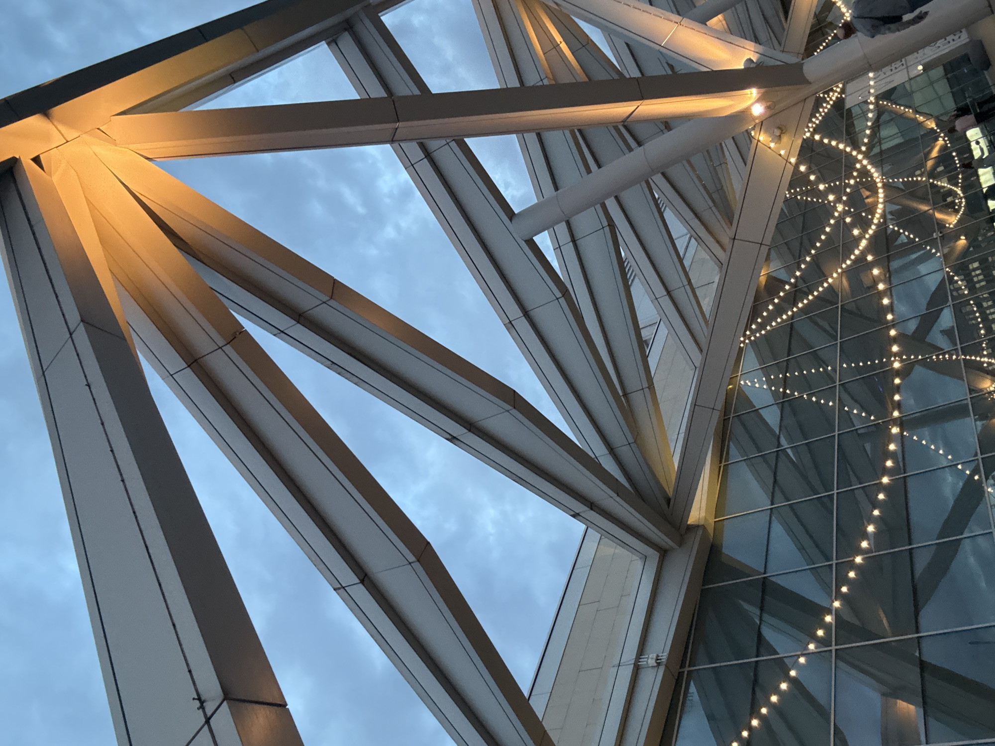 The image depicts a low-angle view of a modern architectural structure in Saint Petersburg, Russia. The primary elements are the steel support beams forming a complex geometric pattern against a cloudy sky. The beams are illuminated, creating a warm, golden glow on their surfaces. Adjacent to the steel structure is a glass facade, decorated with strings of lights arranged in a curved, wave-like pattern. The lighting reflects on the glass. The architectural style is modern, suggesting a contemporary building design. No people are visible in the image. The photograph focuses on the interplay of architecture, lighting, and the natural sky backdrop.