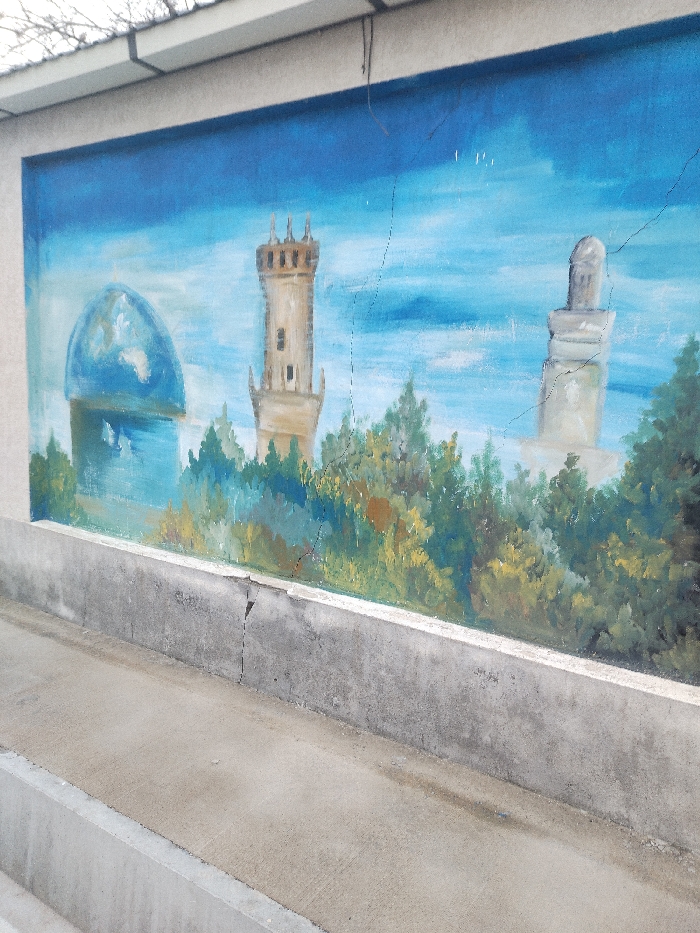 The image displays a concrete wall section in Chighirik, Uzbekistan, featuring a weathered mural. The mural, occupying the central rectangular area of the wall and framed by unpainted light-toned sections above and on the sides, depicts a landscape under a light blue sky with subtle cloud-like formations. Within the mural, from left to right, three prominent structures rise above a dense band of stylized green and brown foliage: a blue domed building featuring a depiction of the Earth, a tan, multi-tiered tower with a crenellated top, and a light grey minaret-like structure with a rounded cap. Multiple fine, dark cracks are visible across the mural's painted surface, some extending into the adjacent unpainted wall. Below the mural, a grey concrete barrier or ledge runs horizontally, presenting a distinct vertical crack near its left-center point. In the immediate foreground, a flat, smooth grey concrete surface serves as a pathway. Above the wall, faint bare tree branches and thin dark cables are visible against an overcast sky.