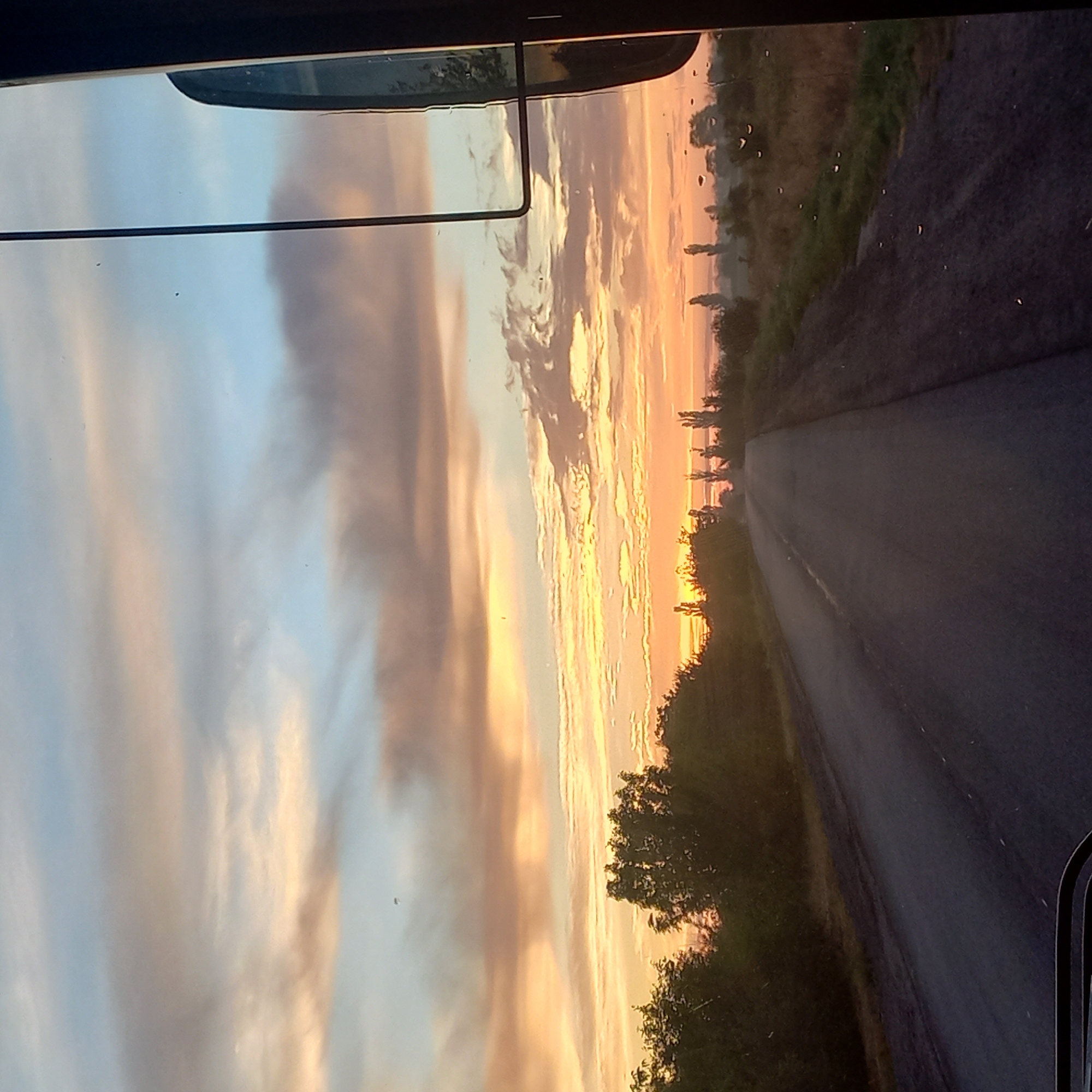 A forward-facing, wide-angle shot captures a road receding into the distance at sunrise. The sky is a gradient of pale blue near the top, transitioning to soft yellows and oranges where the sun is low on the horizon. Wispy, elongated clouds are illuminated by the sun, creating bands of color across the sky. The road itself is asphalt, with a visible white line on the left edge. To the left and right of the road, there are patches of greenery and trees. The scene appears to be on the outskirts of Kropyvnytskyi, Ukraine, as suggested by the rural landscape and the direction of travel. The right side of the frame is partially obscured by a dark, vertical object, possibly a rearview mirror or part of a vehicle's interior structure. Several small, dark spots are visible on the glass, likely water droplets. The overall impression is one of a quiet, early morning journey.