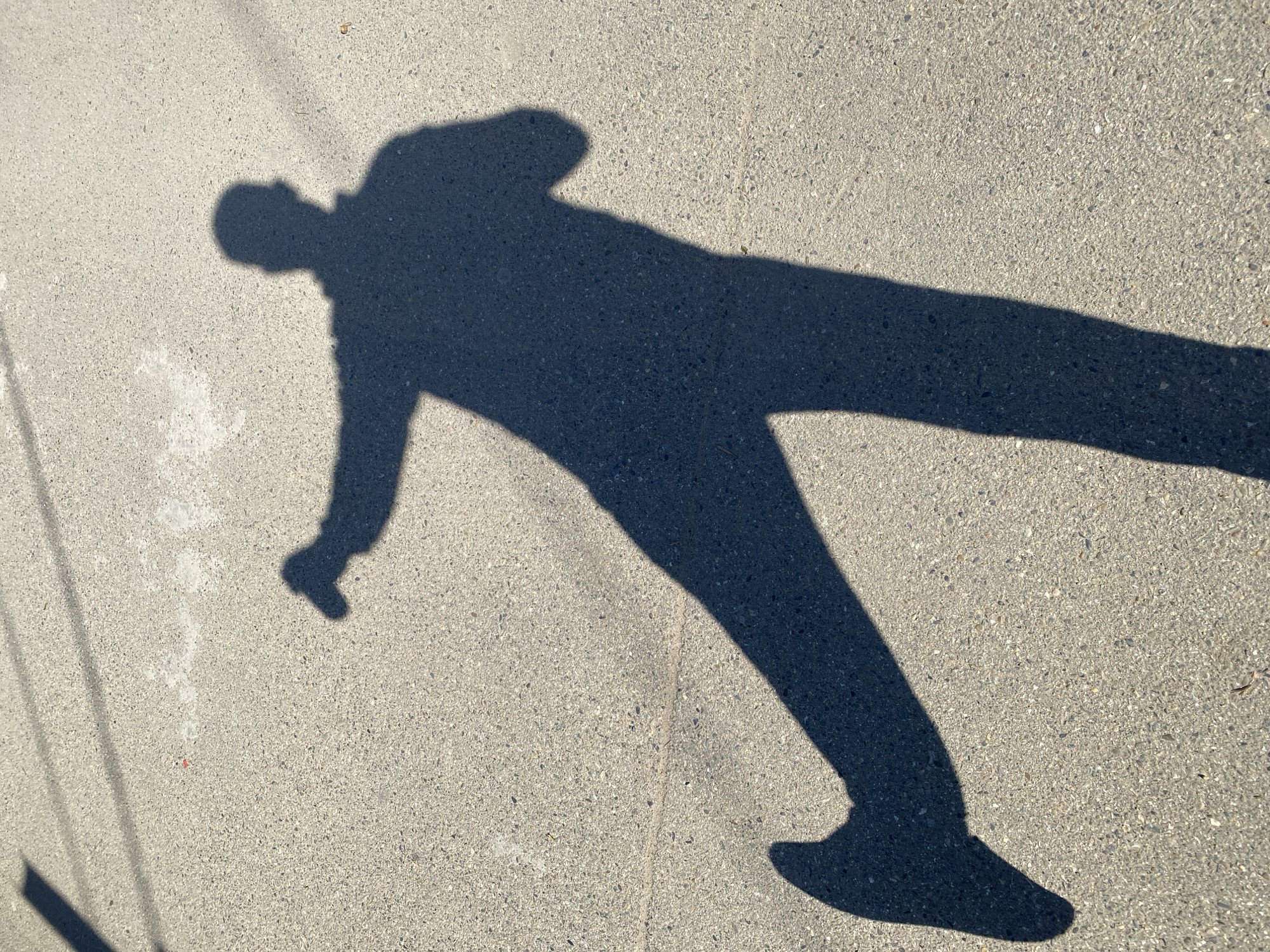 The image displays the long, dark shadow of a person cast distinctly across a light gray, textured paved surface, likely concrete or asphalt. The shadow, occupying the majority of the frame, depicts a dynamic pose with one leg extended far to the right and one arm raised or outstretched, suggesting movement such as stepping, walking, or a playful stance. The head and torso of the shadow are visible towards the upper left.
The sharp definition and strong contrast of the shadow indicate bright, direct sunlight, suggesting the photograph was taken during either the morning or late afternoon when the sun is lower in the sky. In the bottom-left corner of the image, faint, elongated shadows, possibly from overhead wires or poles, are also visible on the pavement. The ground surface is uniform, showing small aggregate particles. There is no discernible text or other prominent objects within the scene, emphasizing the interplay of light and shadow on the outdoor surface.