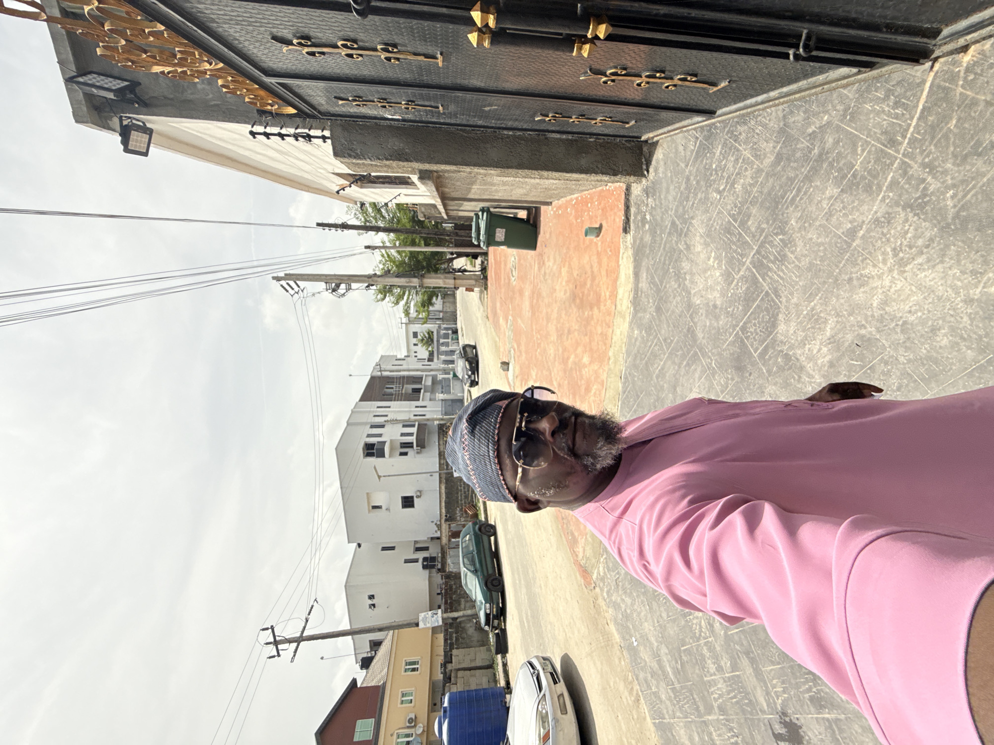 A man is pictured in what appears to be a self-portrait or selfie, presumably in Igboefon, Nigeria. He is wearing a pink shirt, a patterned hat, and eyeglasses. He has a beard. Behind him is a street scene, including buildings, parked cars, utility poles with wires, and a trash receptacle. The road is paved with interlocking concrete pavers. A large ornate metal gate is visible.
