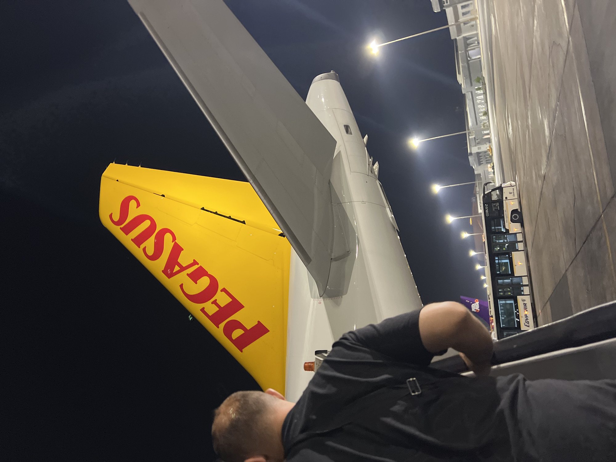 The image shows the rear section of a Pegasus Airlines aircraft at night, with its distinctive yellow tail fin prominently displayed. The aircraft's wing and engine nacelle are also visible. In the foreground, a person viewed from behind, wearing a dark t-shirt and backpack, is standing on boarding stairs, seemingly about to board or disembark. In the background, an EgyptAir bus is parked on the tarmac, with airport buildings and bright floodlights illuminating the scene. The overall impression is of an airport environment at night, likely in Sharm El Sheikh, Egypt, given the presence of EgyptAir branding.