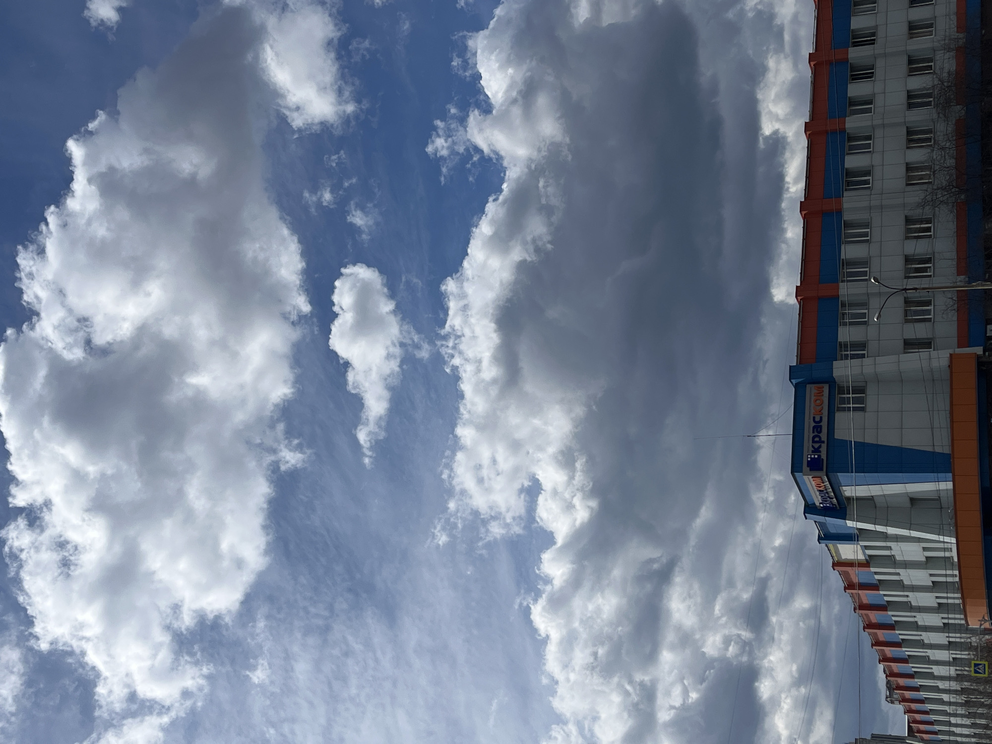 The image, when viewed upright, features a vibrant sky dominating the left side of the frame and a multi-story building on the right. The sky exhibits a mix of clear blue patches and various clouds; some are bright white and fluffy, while others appear denser and darker grey. This suggests a dynamic and partly cloudy weather condition during daytime.
On the right, a modern building rises, characterized by its light grey or white paneling. It features distinct vertical and horizontal accents in bright blue and orange, adding to its contemporary aesthetic. Numerous rectangular windows are visible across its facade. A street lamp, featuring two light fixtures, stands in front of the building. Behind the lamp, the skeletal branches of a deciduous tree, devoid of leaves, are visible, suggesting a time of year such as late autumn, winter, or early spring.
Prominently displayed on the building's facade is a blue sign with white Cyrillic text, which reads "КРАСКОМ" (KRASKOM). The scene is static, capturing a moment of an urban landscape in Krasnoyarsk, Russia.