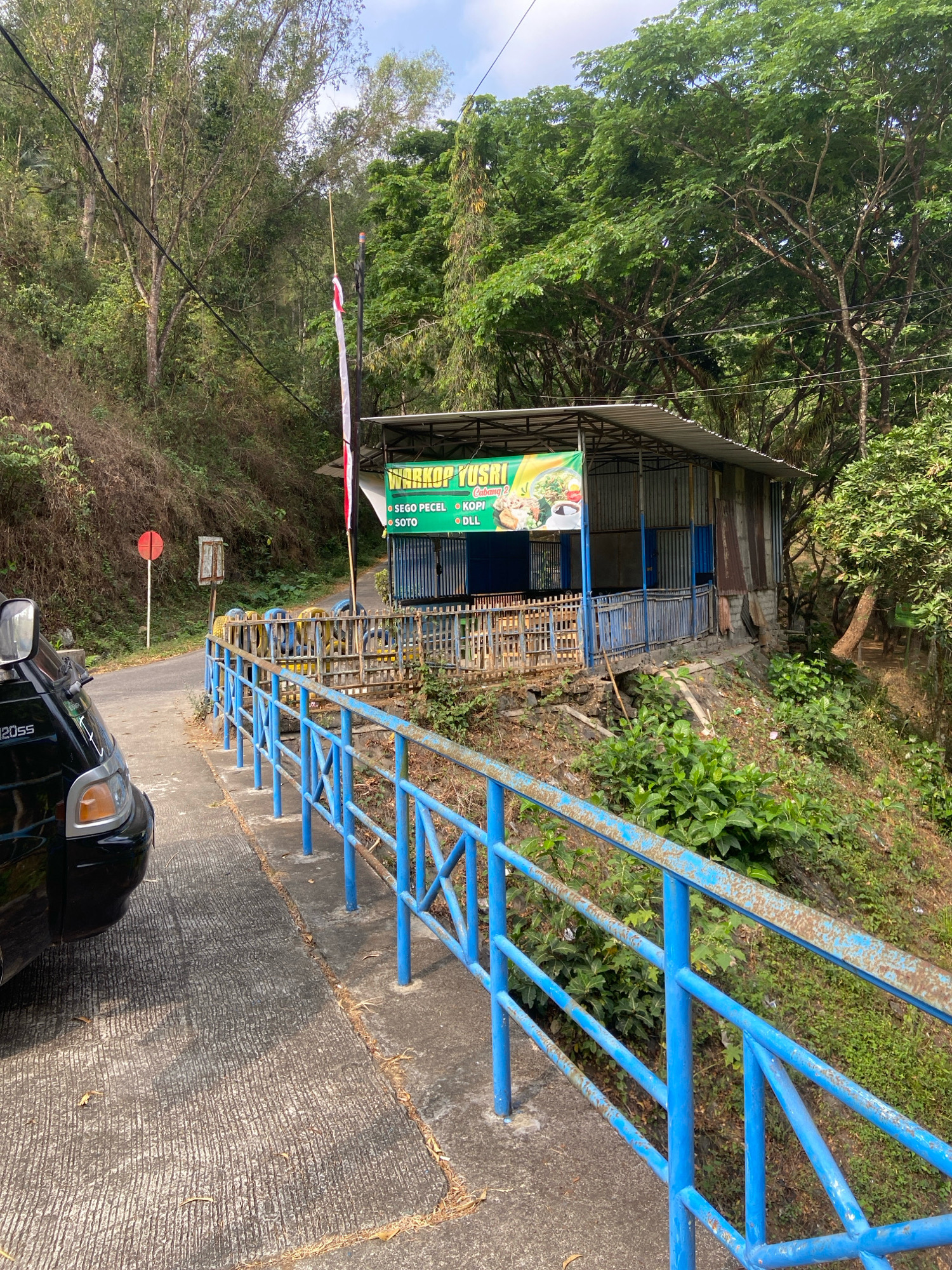 A roadside warung (food stall) named "WARKOP YUSRI CABANG 2" is situated on a sloped hillside overlooking a road. The warung has a metal roof and corrugated metal walls, with a blue metal railing in front of it. A green banner above the warung displays food items like "SEGO PECEL", "KOPI", and "SOTO". A red and white Indonesian flag flies on a pole near the warung. A black vehicle is parked on the concrete road to the left of the scene. The surrounding environment is lush with green trees and vegetation. The image appears to be taken in Segawe, Indonesia.