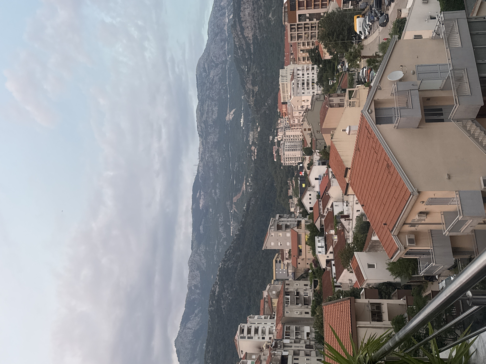 The image presents a high-angle view of a residential area in Budva, Montenegro, with a range of buildings nestled against a backdrop of lush green hills and rugged mountains under a cloudy sky. In the foreground, several multi-story buildings with red-tiled roofs dominate the scene. These buildings feature balconies, windows, and external air conditioning units, indicative of residential apartments or homes. Some of these structures are painted in light colors like beige and white, with some displaying more complex architectural designs.
Further into the scene, taller, modern apartment blocks with numerous windows and balconies are visible, interspersed with smaller houses. The architecture suggests a mix of older and newer constructions. The streets are narrow and winding, with cars parked along the roadside. One yellow vehicle, possibly a van, is notable among the parked cars. The presence of palm trees and other greenery suggests a Mediterranean climate.
In the background, the landscape rises dramatically with densely forested hills leading up to barren, rocky mountains. A road or path can be seen cutting across one of the mountainsides, with evidence of some construction or clearing. The sky is overcast with a blend of white and gray clouds, creating a diffused light that softens the overall scene. There are no people clearly visible in the image, but the presence of cars and parked vehicles implies human activity and habitation. The image captures a dense urban environment seamlessly integrated with the natural mountainous terrain.