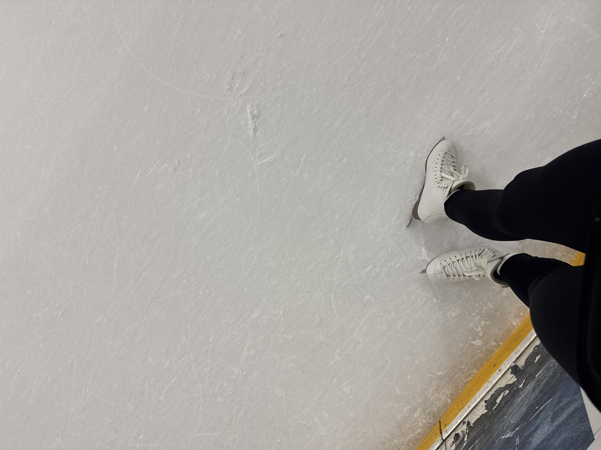 The image depicts a pair of white ice skates on a scratched ice rink. The skater is wearing black leggings. The bottom right corner shows part of the rink's barrier, featuring a yellow stripe and a darker section. The location appears to be an indoor ice rink, but the city of Tbilisi, Georgia cannot be confirmed solely from the visual information provided.