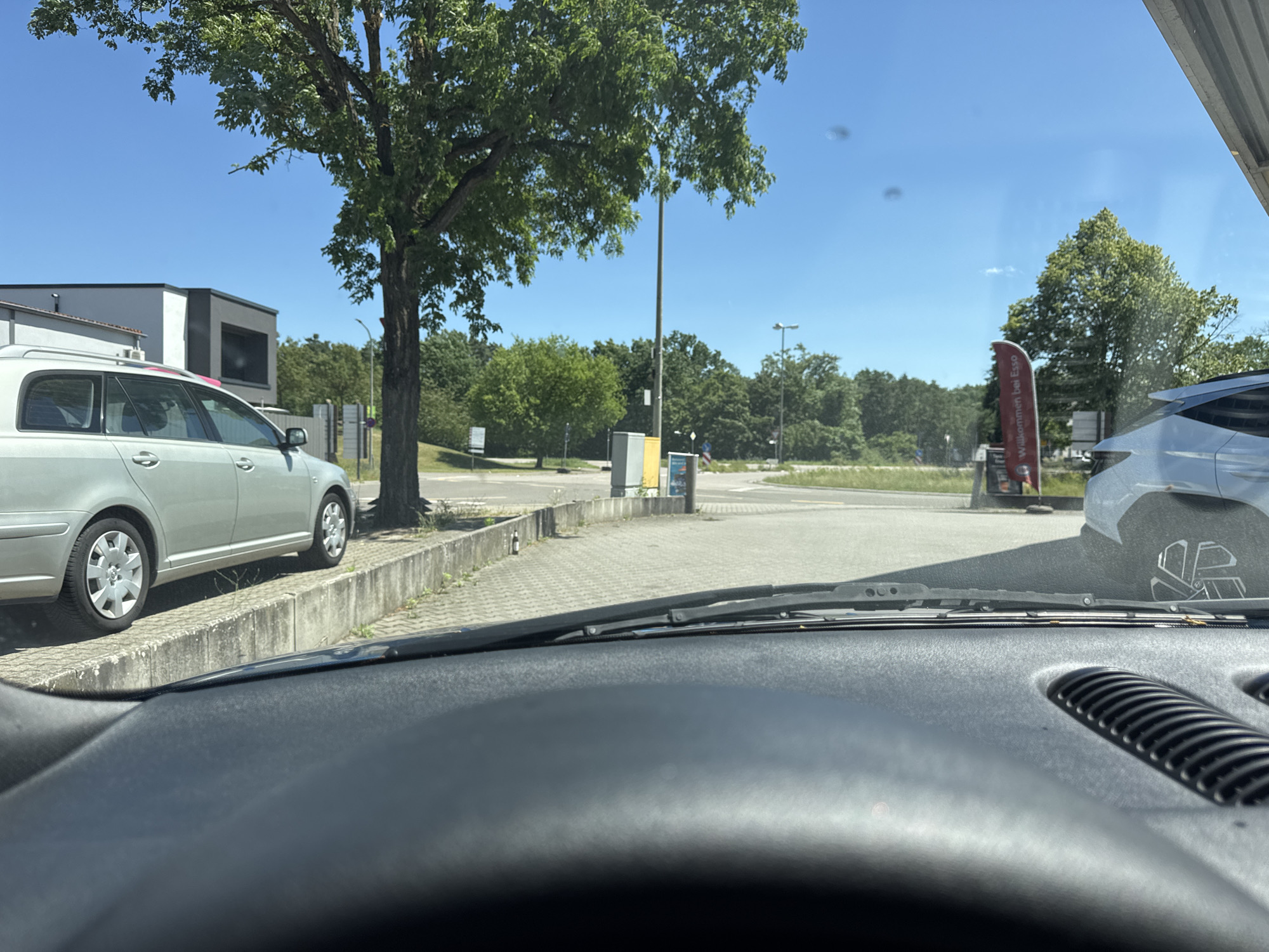 The image, taken from the interior of a vehicle looking forward through the windshield, depicts an outdoor scene on a sunny day, likely in the vicinity of a gas station. In the foreground, the dark dashboard of the car is visible. Beyond the windshield, a light-colored station wagon is parked on the left, parallel to a concrete curb and a paved area. Further to the right, another car, predominantly white, is visible from the rear quarter, its wheel and part of its body visible.
A prominent tree with green foliage stands to the left of center, its trunk thick and dark. Behind the station wagon, a modern building with white and dark grey elements is partially visible. In the middle ground, a street or parking lot extends, with a red and white flag banner standing on the right side, which appears to read "Willkommen bei Esso." Several trash receptacles, one yellow and one grey, are placed along the edge of the paved area. In the background, dense green trees form a treeline under a clear blue sky, with lampposts visible in the distance.
The location is identified as being in the city of Bellheim, Germany, likely at a service station due to the "Esso" signage. There are no people or animals clearly visible in the image. The scene appears to be static, with vehicles parked and no overt activity taking place.