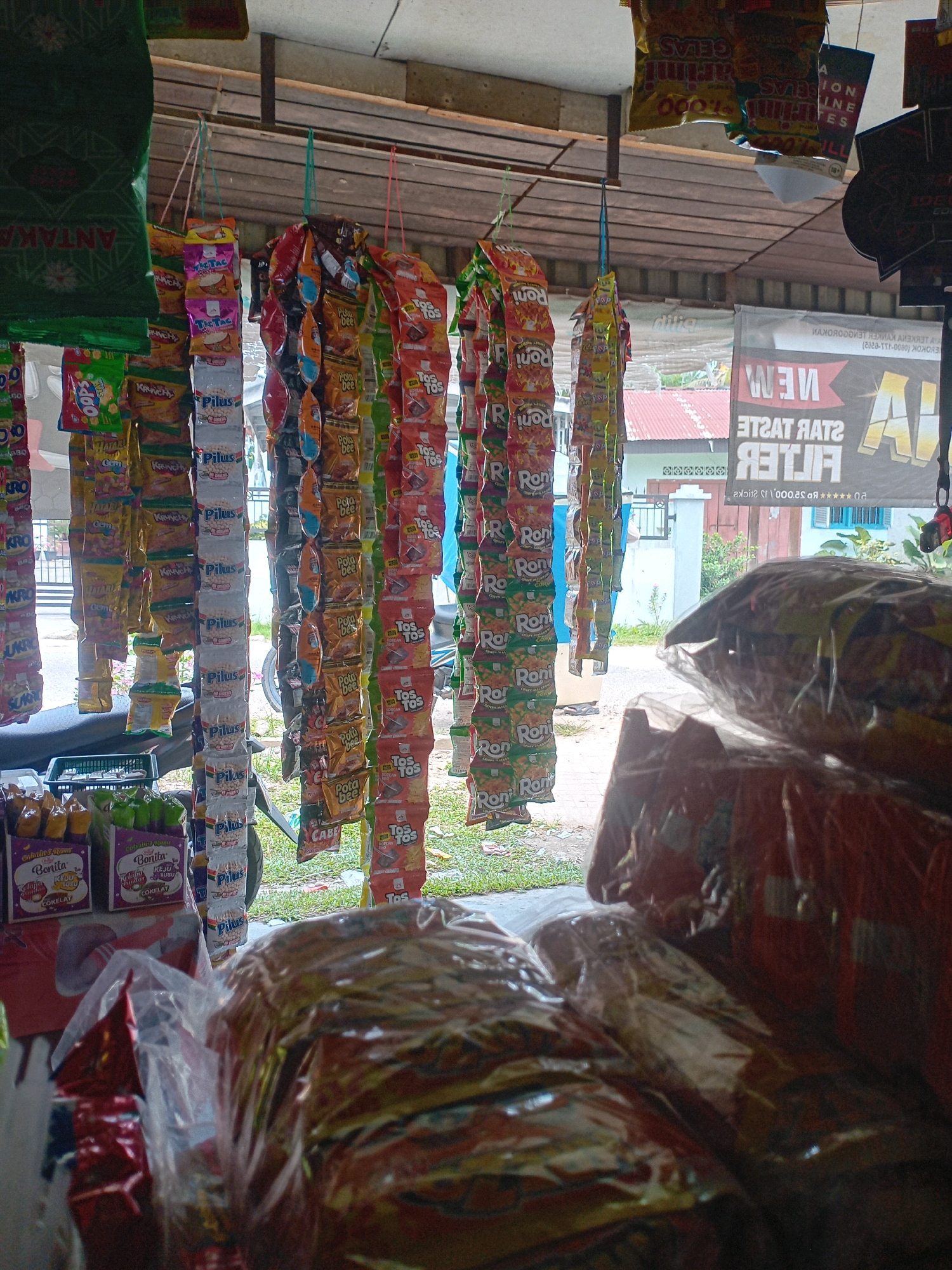 The image depicts the interior of a small roadside stall or convenience store in Pekanbaru, Indonesia, likely a warung kecil. The foreground is dominated by stacks of bagged snacks and goods, some wrapped in transparent plastic. Hanging from a rack above are numerous small snack packets, arranged in vertical rows. These packets display colorful branding, including the recognizable logos of "TOS TOS" and "Pila Bee". Other brands like "Pilus" and "Roni" are also visible on the hanging snacks.
In the background, through an opening or window of the stall, a residential area with red-tiled roofs and some greenery is visible, typical of urban or suburban Indonesian landscapes. A large banner with text in Indonesian, including "STAR RATE" and "FILTER," is mounted on the wall behind the hanging snacks. The overall impression is of a modest, well-stocked retail space catering to everyday needs. There are no people visible in the frame. The lighting suggests it is daytime, possibly with some overhead artificial lighting from the stall's roof.